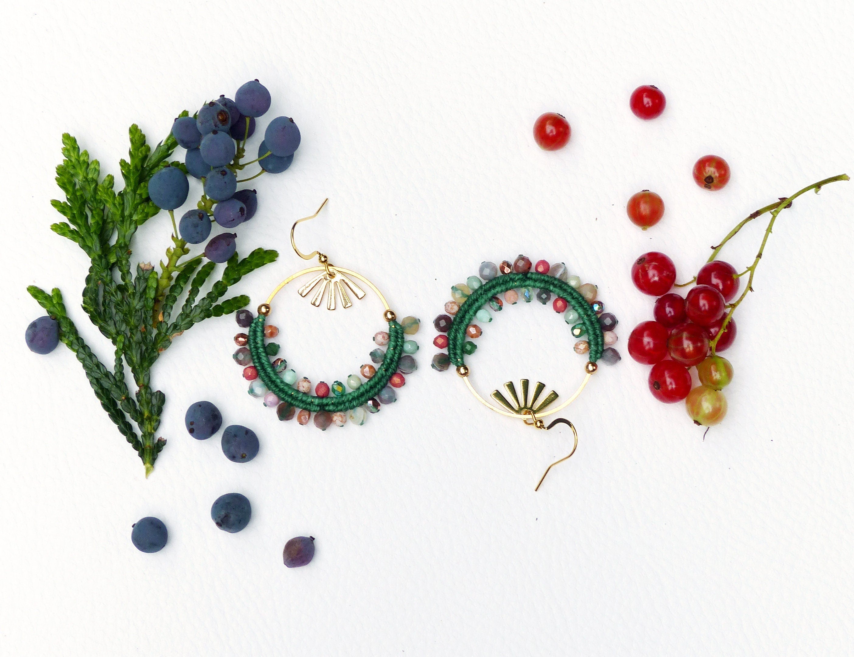 Peut inclure: Une paire de boucles d'oreilles créoles vertes et dorées avec des perles colorées et un motif en forme d'éventail. Les boucles d'oreilles sont photographiées sur une surface blanche avec des baies et de la verdure.
