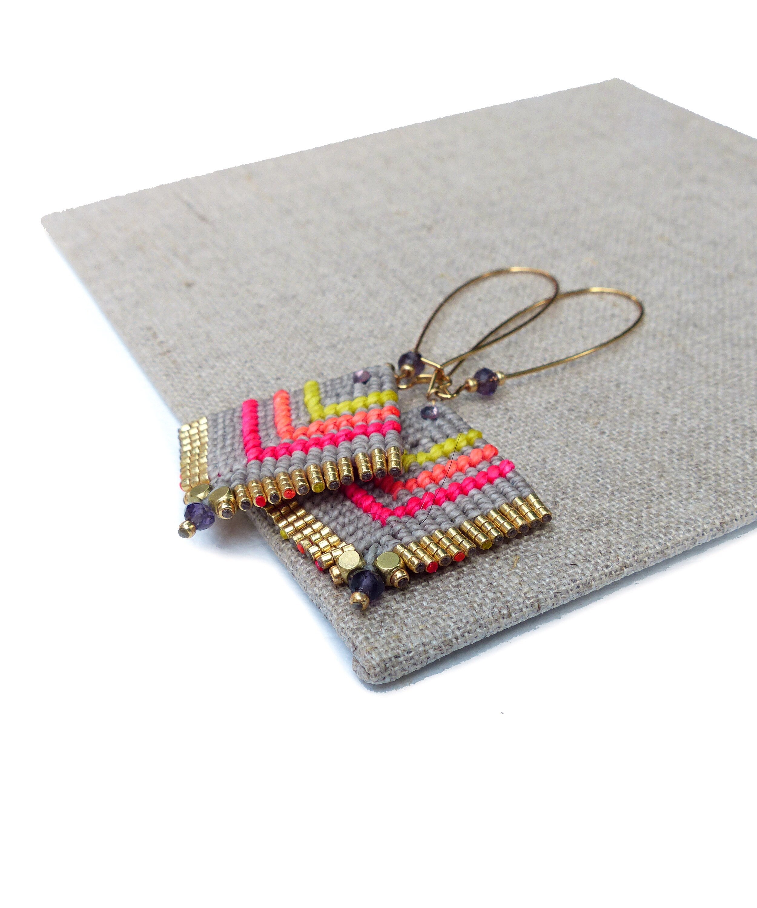 Peut inclure: Une paire de boucles d'oreilles géométriques avec un motif gris, rose et jaune. Les boucles d'oreilles sont faites de petites perles et ont des accents dorés.