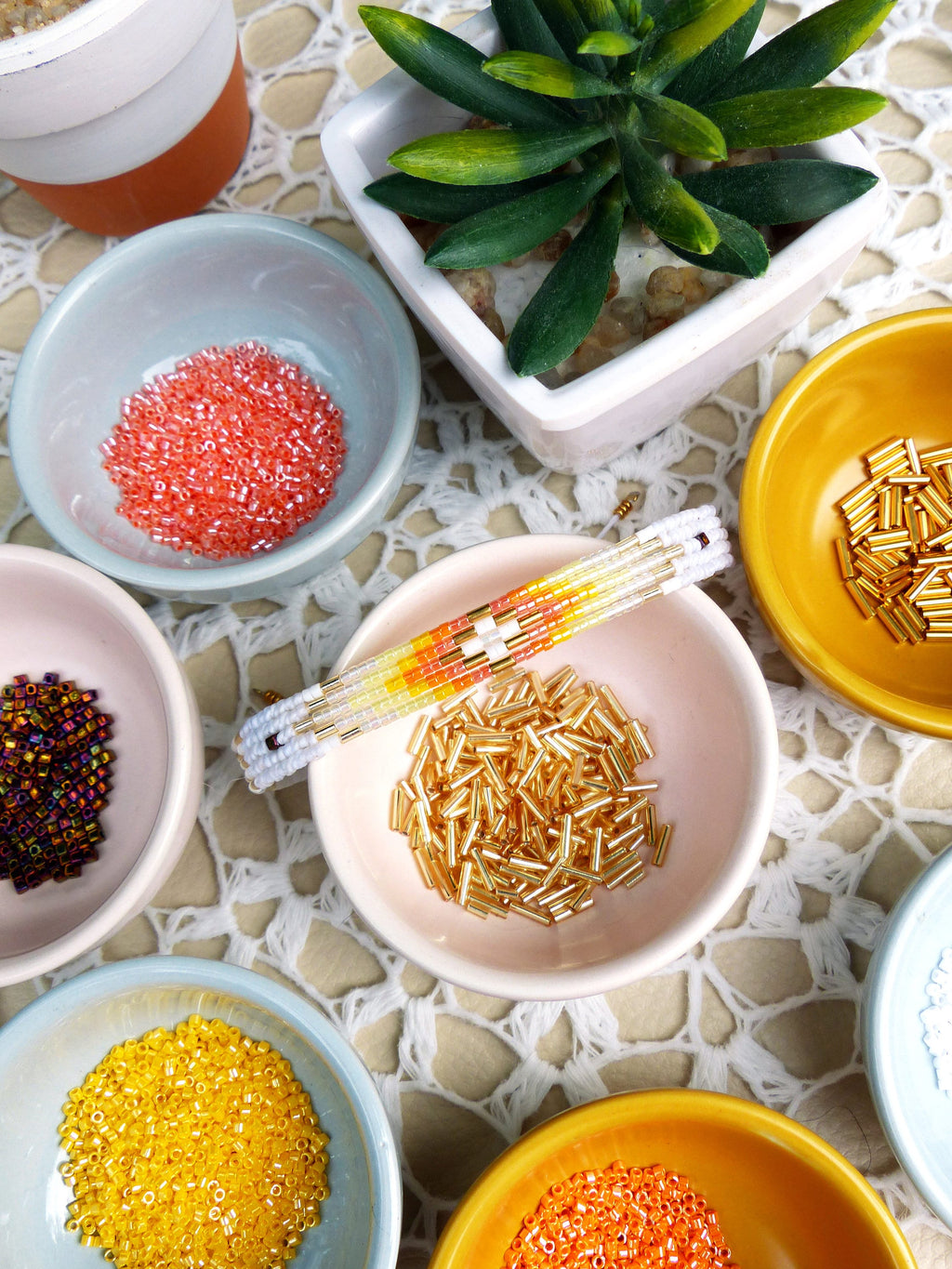 Peut inclure: Assortiment de perles de rocaille colorées dans de petits bols, y compris orange, jaune, or et irisé. Un bracelet en perles avec des perles orange, jaunes et blanches est posé sur un bol de perles dorées.