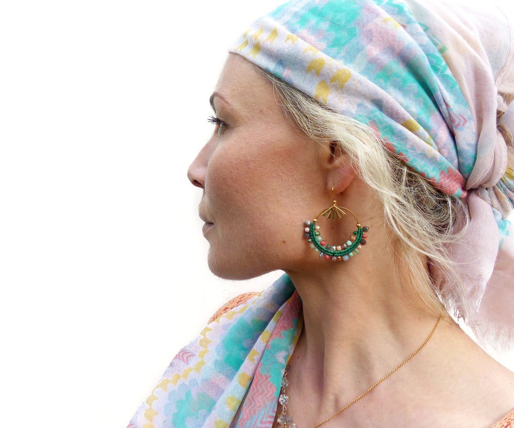 Peut inclure: Une femme portant un foulard à motifs turquoise et rose pâle avec des boucles d'oreilles dorées ornées de perles vertes et multicolores. Les boucles d'oreilles sont en forme de cerceau avec un motif soleil doré.