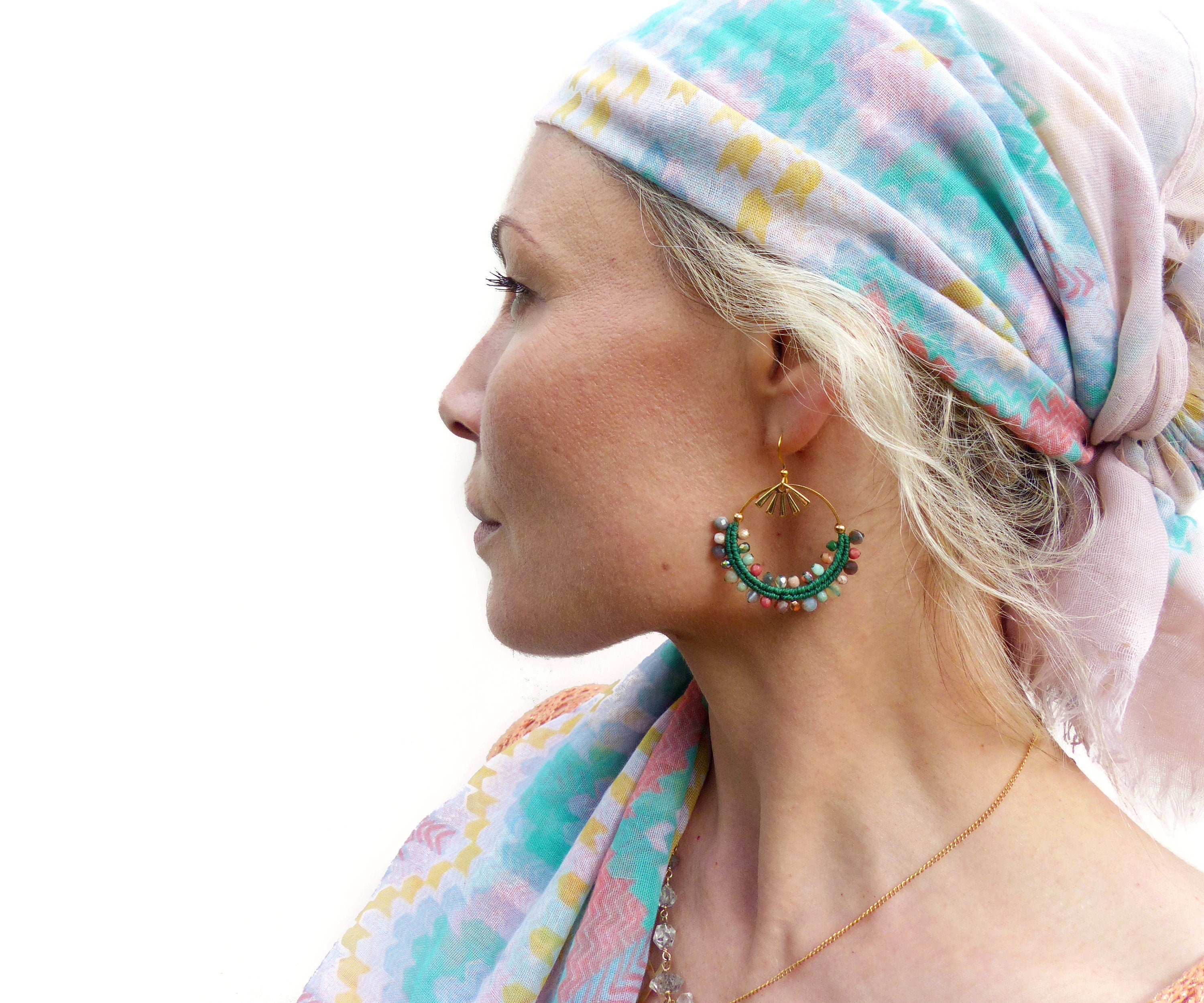 Peut inclure: Une femme portant un foulard à motifs turquoise et rose pâle avec des boucles d'oreilles dorées ornées de perles vertes et multicolores. Les boucles d'oreilles sont en forme de cerceau avec un motif soleil doré.