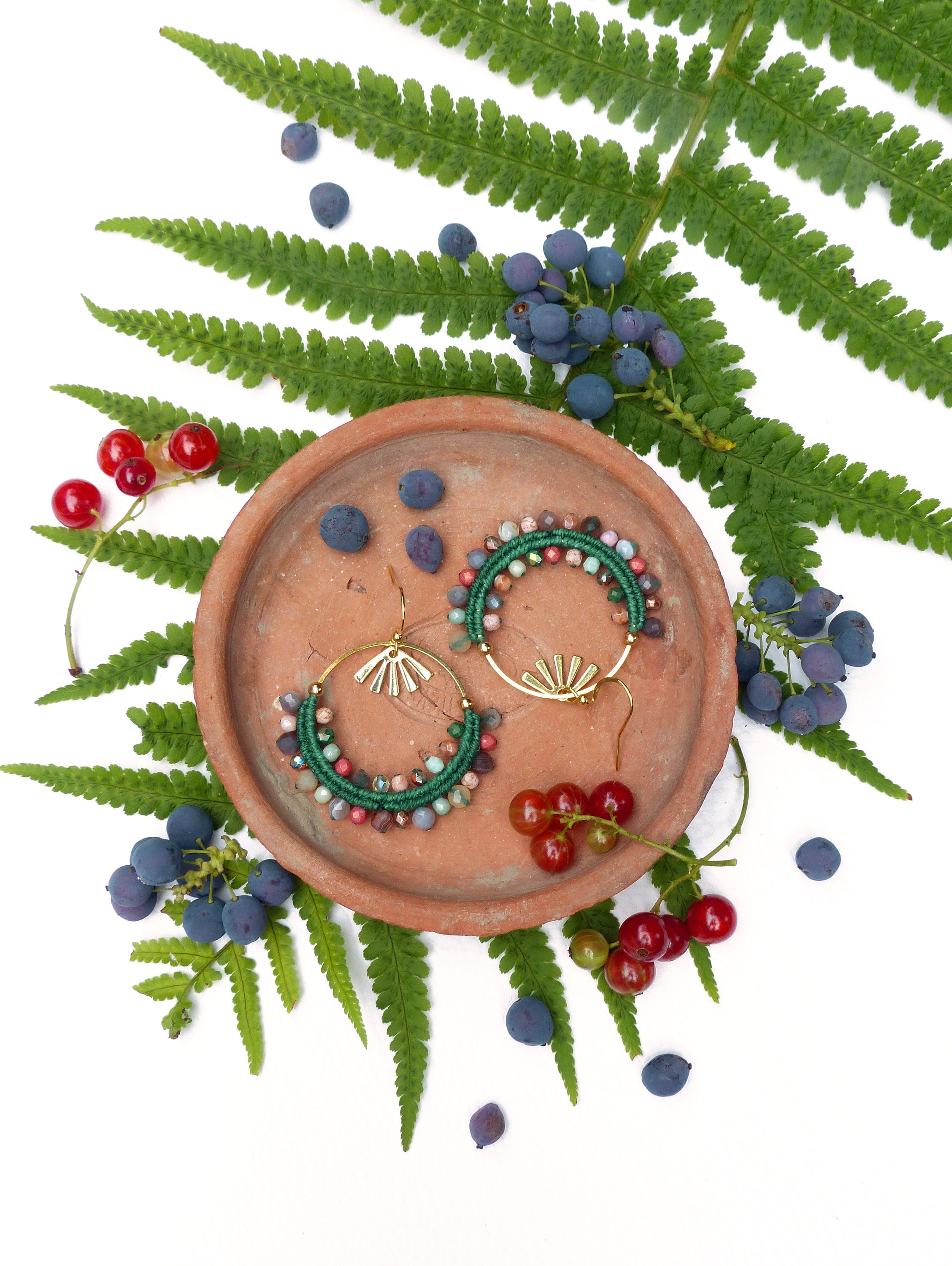 Peut inclure: Une paire de boucles d'oreilles créoles en perles vertes et brunes avec des accents dorés sur une assiette en terre cuite entourée de fougères vertes et de baies rouges et bleues.