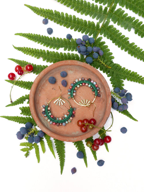Peut inclure: Une paire de boucles d'oreilles créoles en perles vertes et brunes avec des accents dorés sur une assiette en terre cuite entourée de fougères vertes et de baies rouges et bleues.