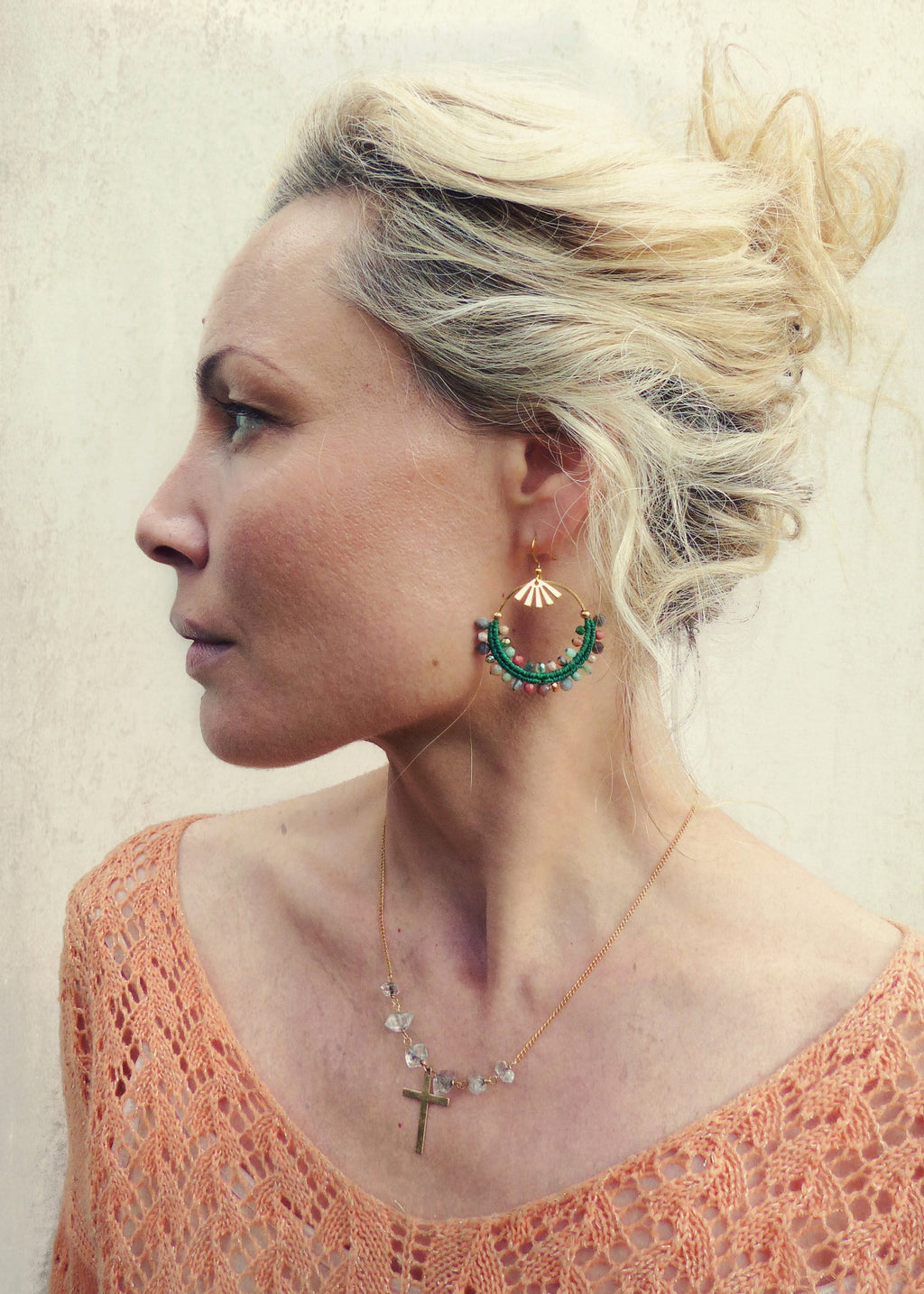 Peut inclure: Une femme portant un pull en crochet couleur pêche avec un collier en chaîne dorée orné d'une petite croix et de perles de cristal claires. Elle porte de grandes boucles d'oreilles créoles avec des perles vertes et roses et un motif soleil doré.