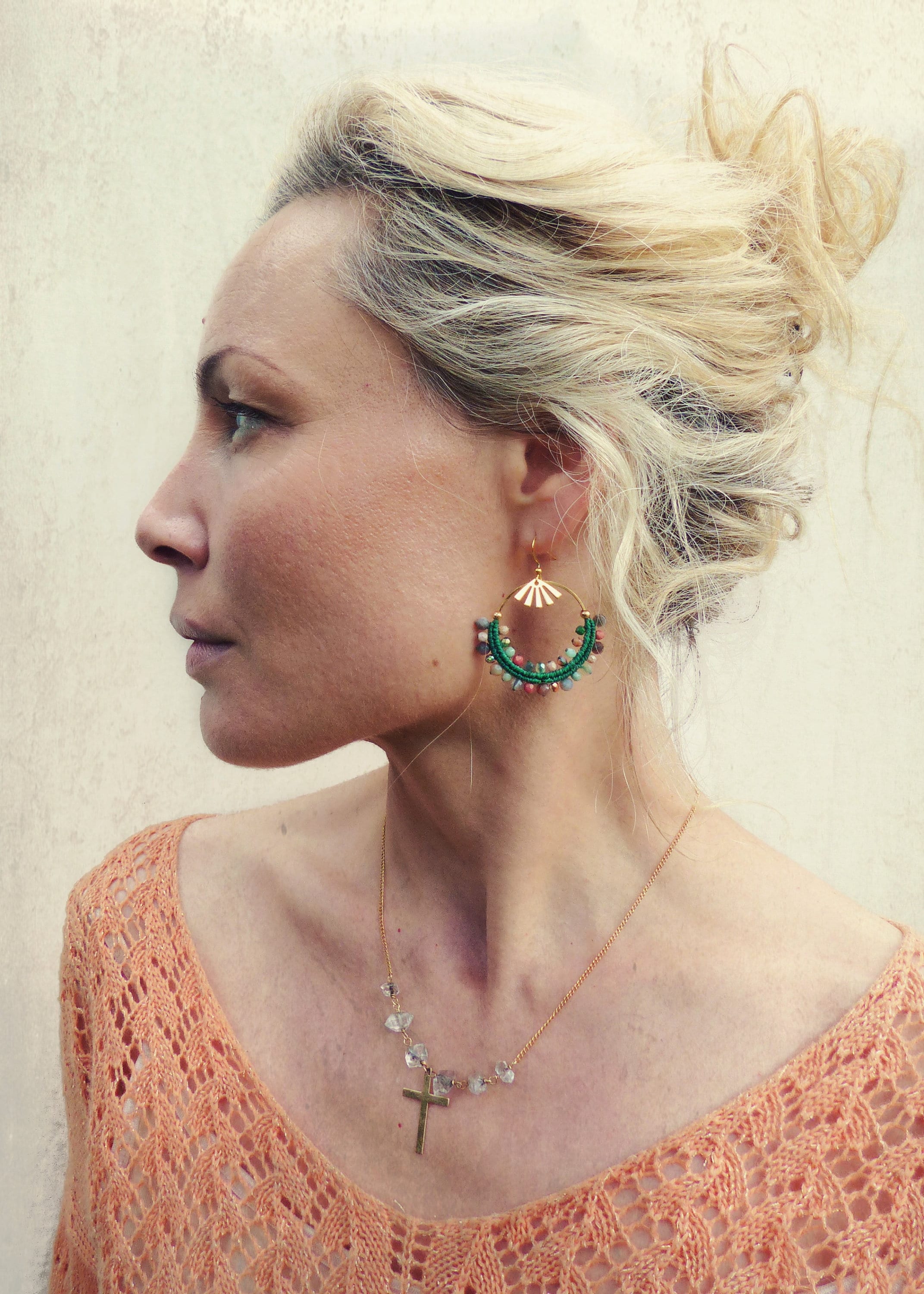 Peut inclure: Une femme portant un pull en crochet couleur pêche avec un collier en chaîne dorée orné d'une petite croix et de perles de cristal claires. Elle porte de grandes boucles d'oreilles créoles avec des perles vertes et roses et un motif soleil doré.