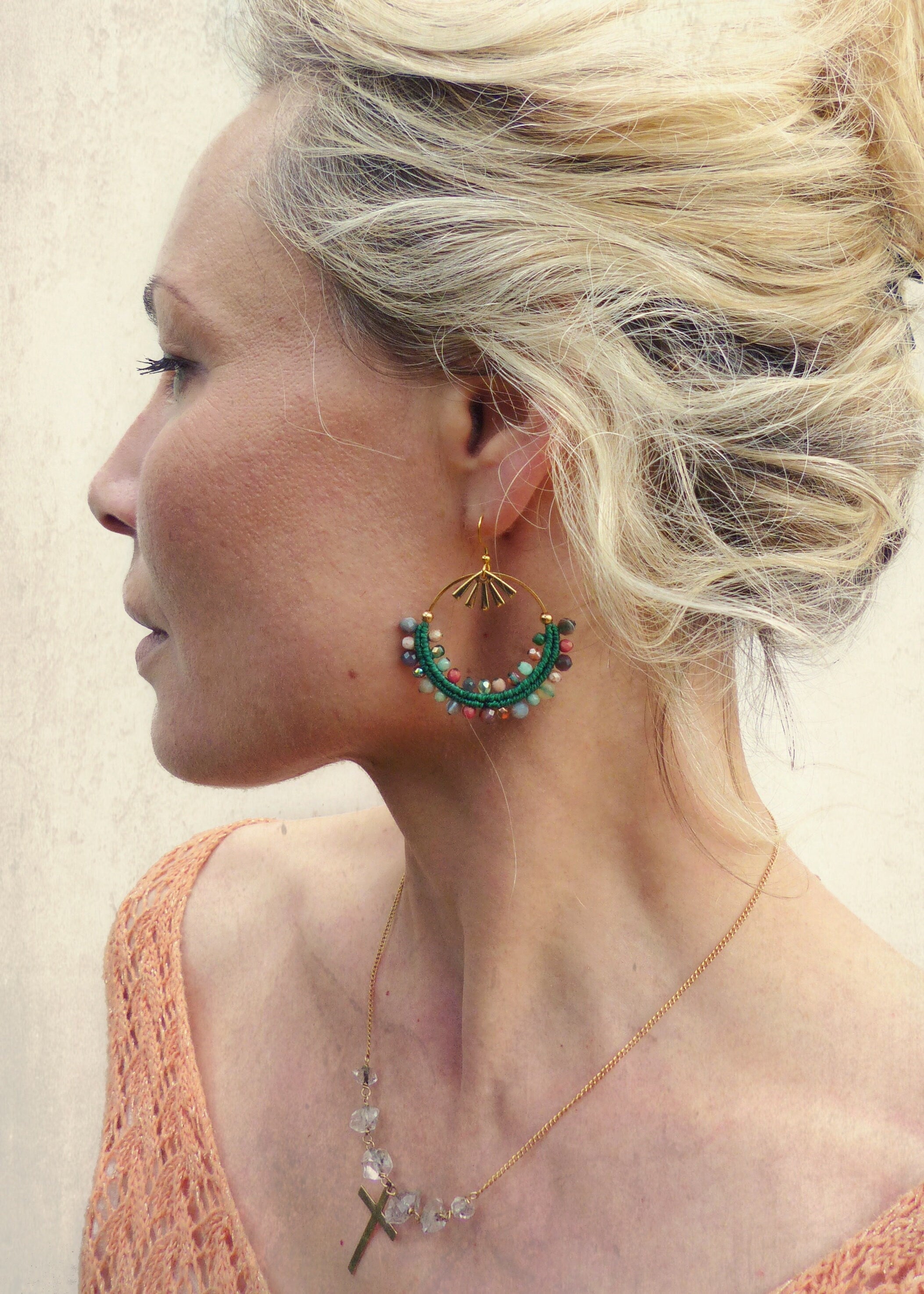 Peut inclure: Une femme portant un collier en or avec un pendentif en forme de croix et des perles de cristal claires. Elle porte également une paire de boucles d'oreilles créoles en or avec des perles vertes et multicolores.
