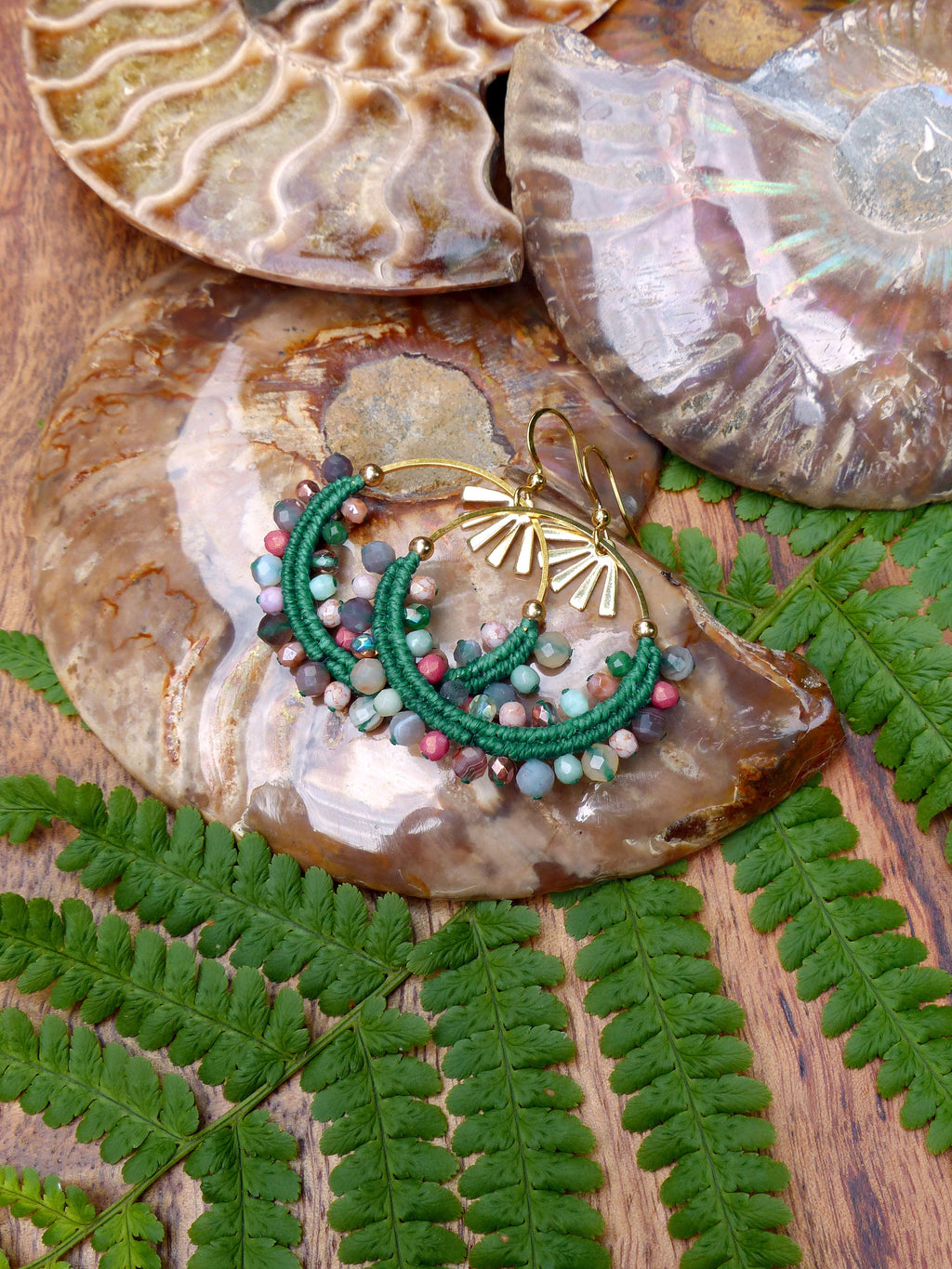 Peut inclure: Une paire de boucles d'oreilles en perles vertes et roses avec des cercles dorés et un motif soleil. Les boucles d'oreilles sont présentées sur une coquille fossilisée brune et blanche.