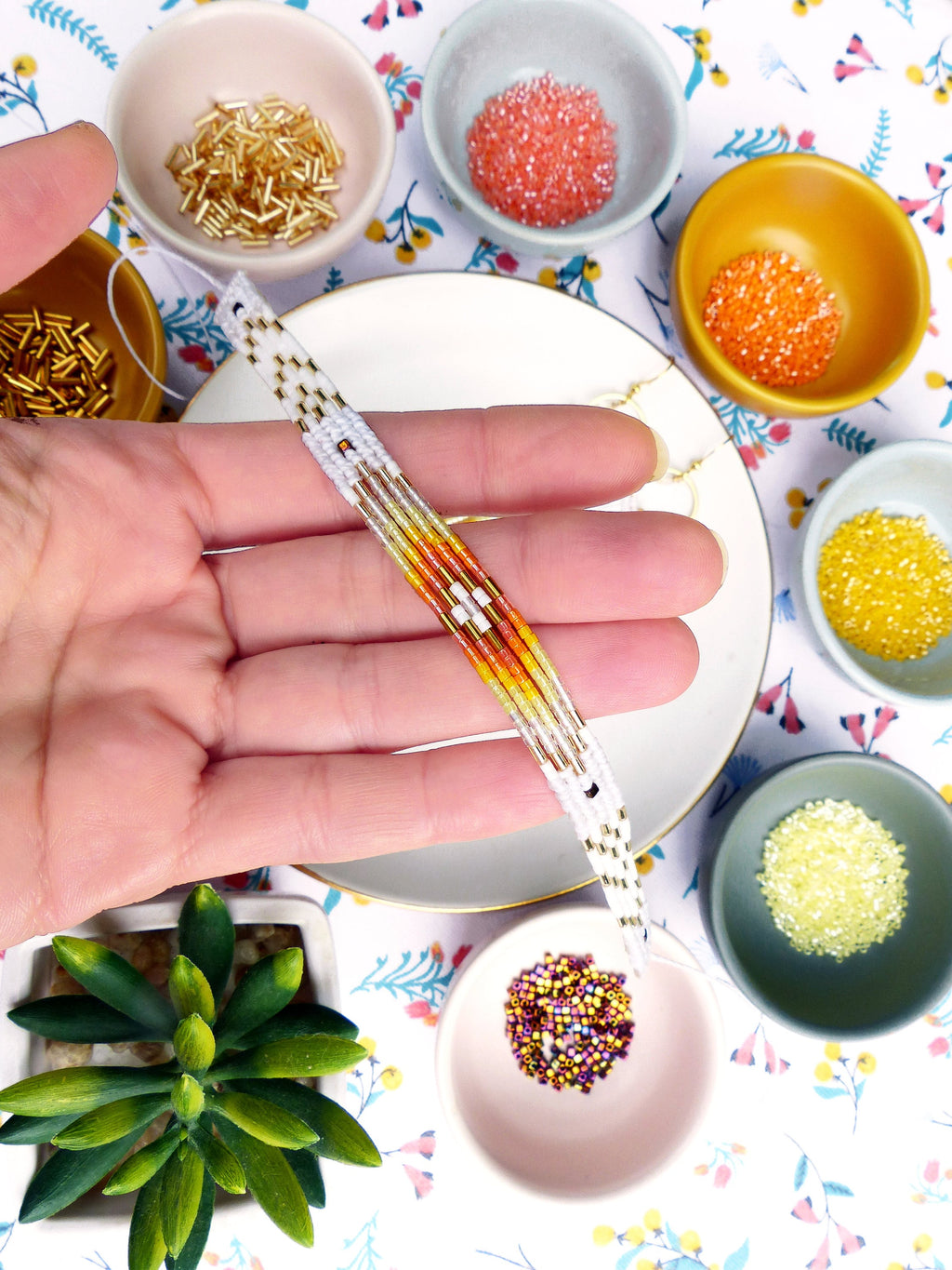 Peut inclure: Gros plan sur une main tenant un bracelet en perles avec un motif dégradé coloré. Le bracelet est composé de perles blanches, orange, jaunes et dorées. L'image montre également plusieurs petits bols remplis de perles de différentes couleurs.