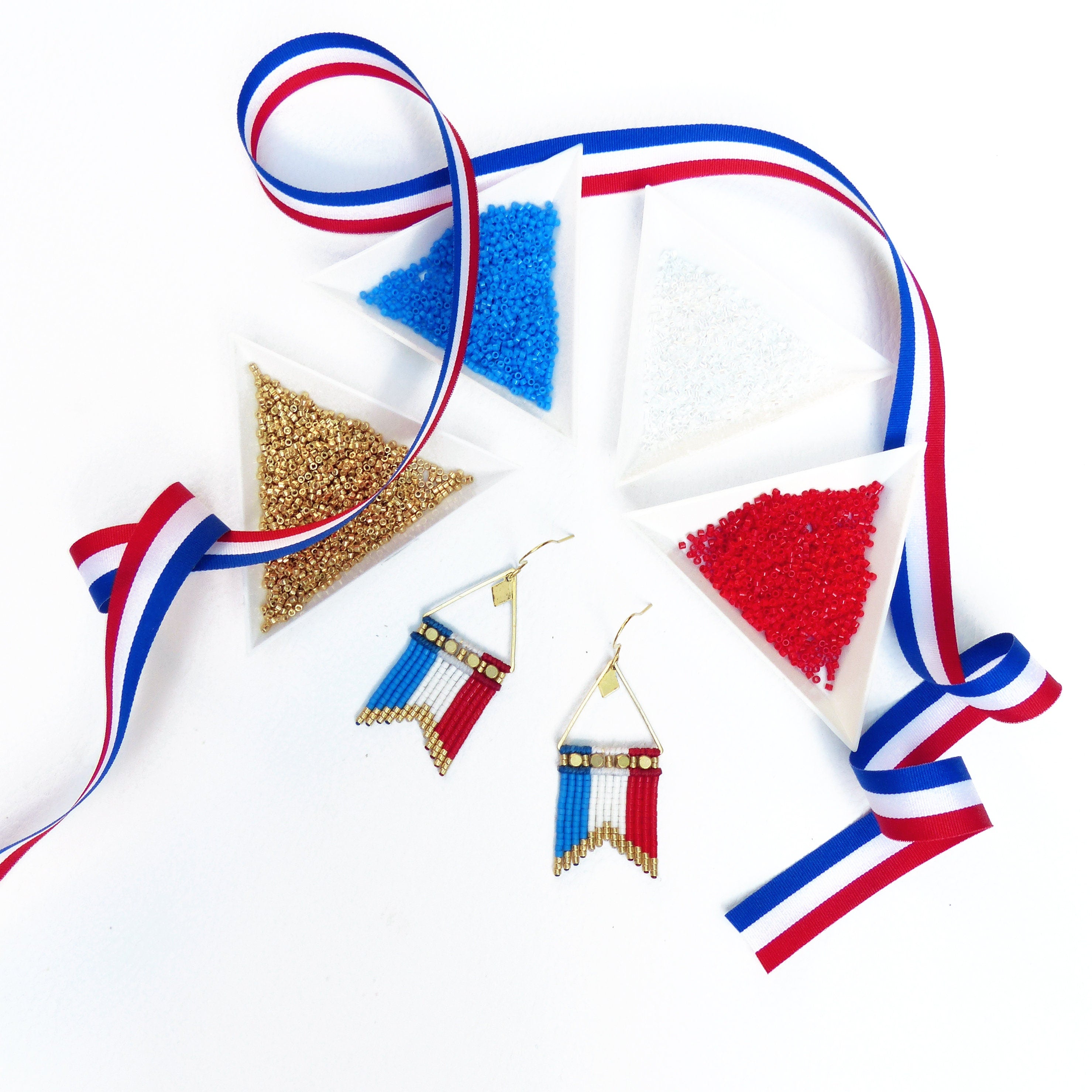 Peut inclure: Boucles d'oreilles faites à la main avec un motif de drapeau, composées de perles bleues, blanches et rouges. Les boucles d'oreilles sont présentées avec de petits contenants triangulaires de perles bleues, blanches, rouges et dorées, et un ruban tricolore.