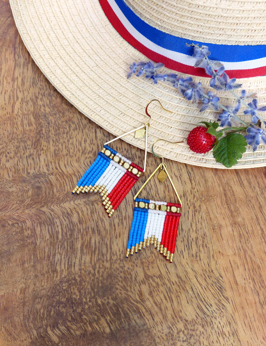Peut inclure: Boucles d'oreilles triangulaires dorées avec un motif de drapeau en perles rouges, blanches et bleues. Elles sont posées sur une surface en bois avec un chapeau de paille, des brins de lavande et une baie rouge.