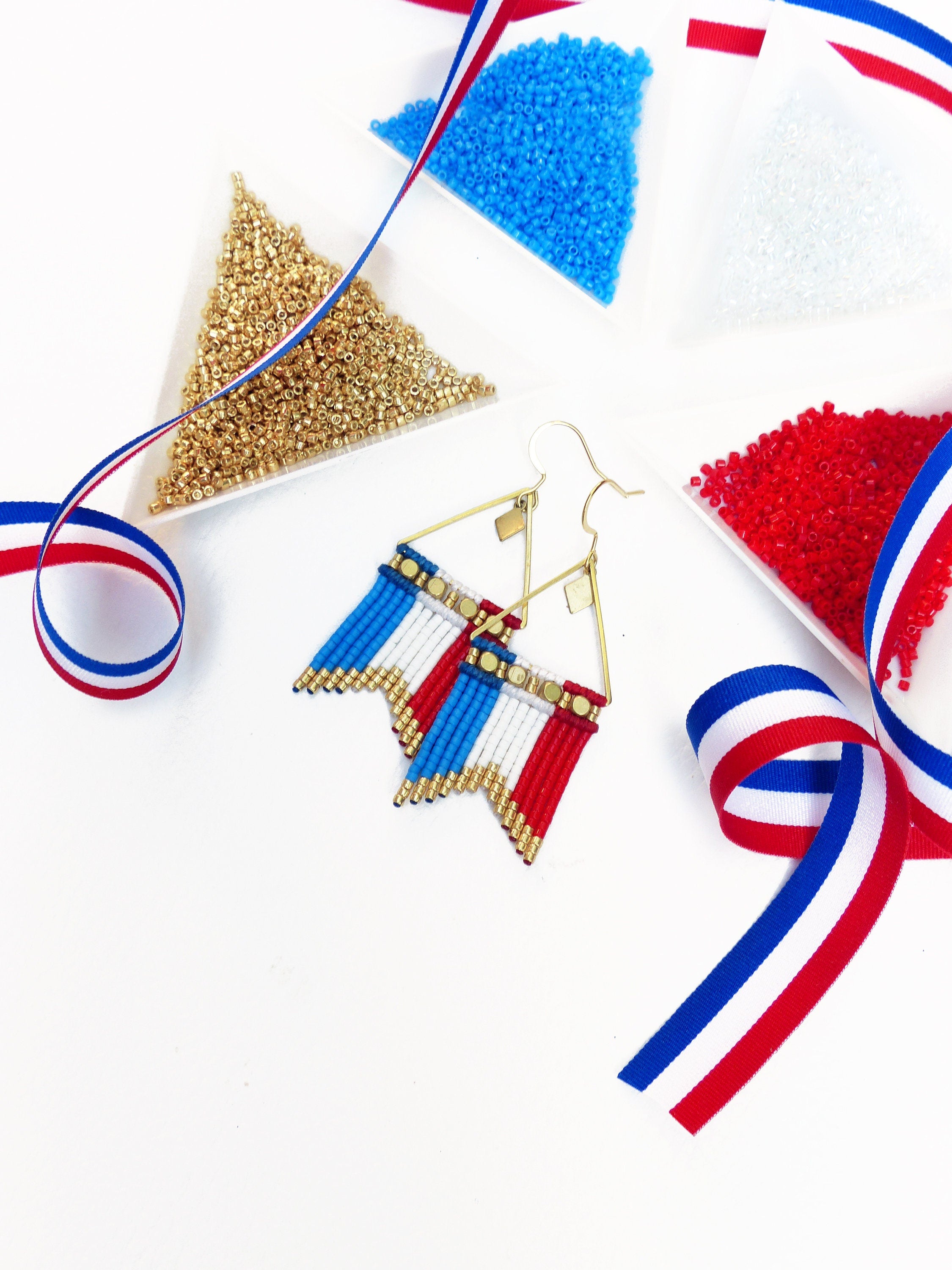 Peut inclure: Boucles d'oreilles faites à la main avec un motif de drapeau, composées de perles bleues, blanches et rouges. Les boucles d'oreilles ont un cadre géométrique doré. L'image montre également des perles dorées, bleues, blanches et rouges dans des récipients triangulaires, ainsi qu'un ruban tricolore.