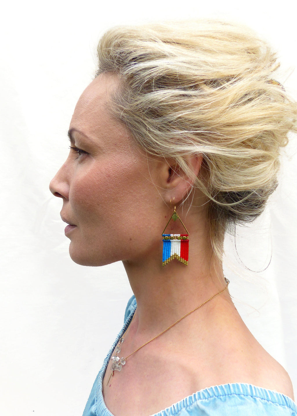 Peut inclure: Boucles d'oreilles triangulaires dorées avec un motif de drapeau en perles rouges, blanches et bleues. Les boucles d'oreilles sont suspendues à des crochets dorés. Un collier en or avec un pendentif est également visible.