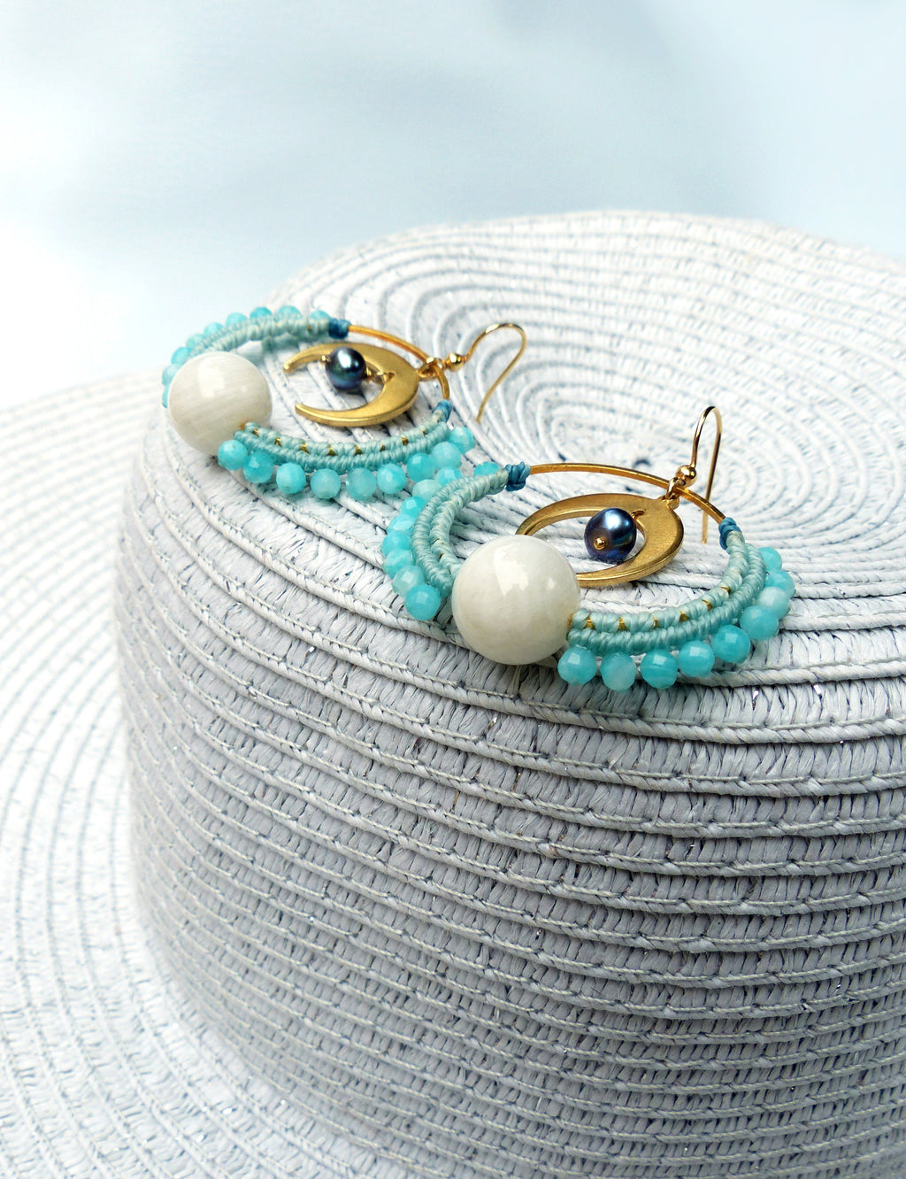Peut inclure: Une paire de boucles d'oreilles créoles dorées avec des perles bleues et blanches et un charme en forme de croissant de lune. Les boucles d'oreilles sont sur un chapeau de paille blanc.
