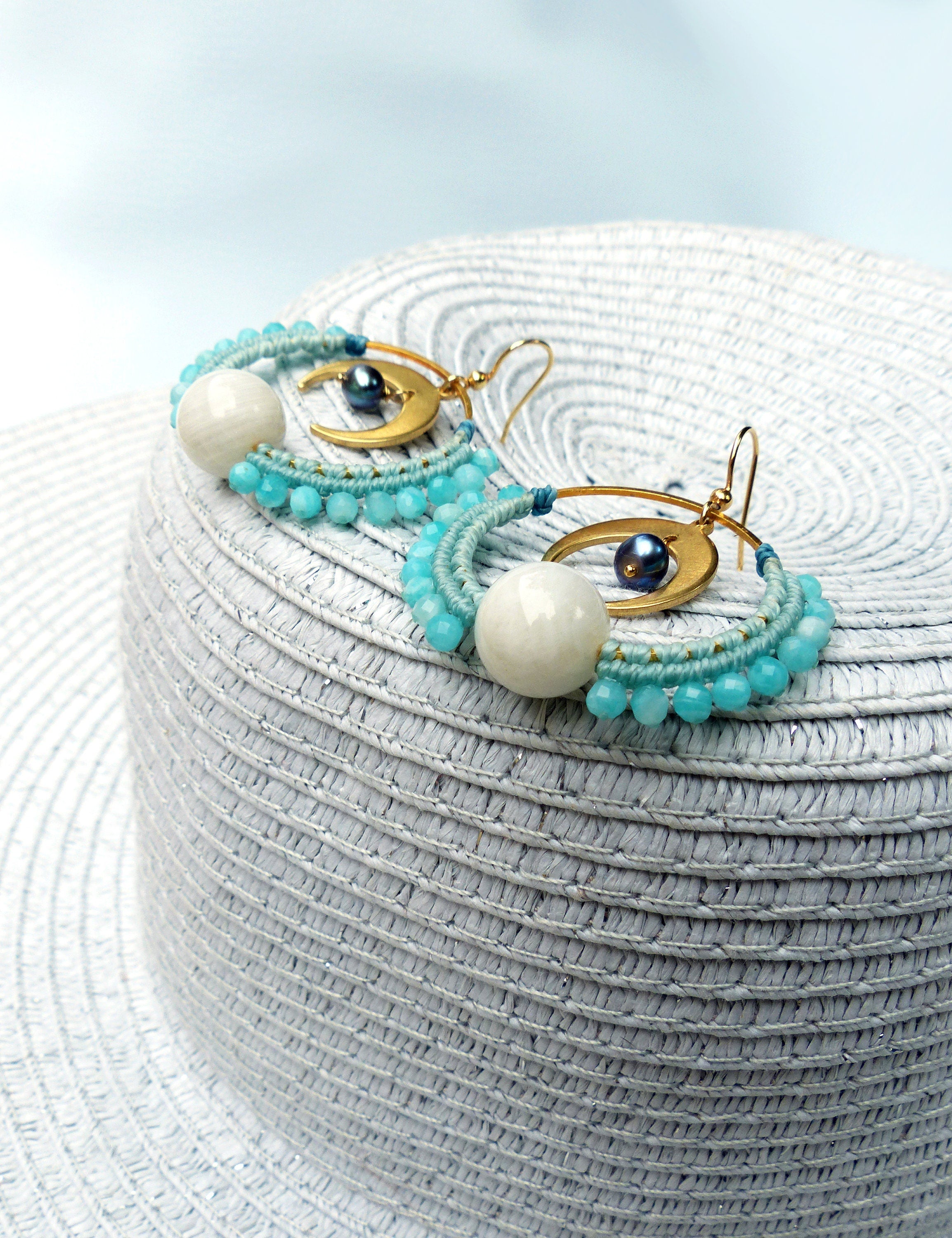 Peut inclure: Une paire de boucles d'oreilles créoles dorées avec des perles bleues et blanches et un charme en forme de croissant de lune. Les boucles d'oreilles sont sur un chapeau de paille blanc.
