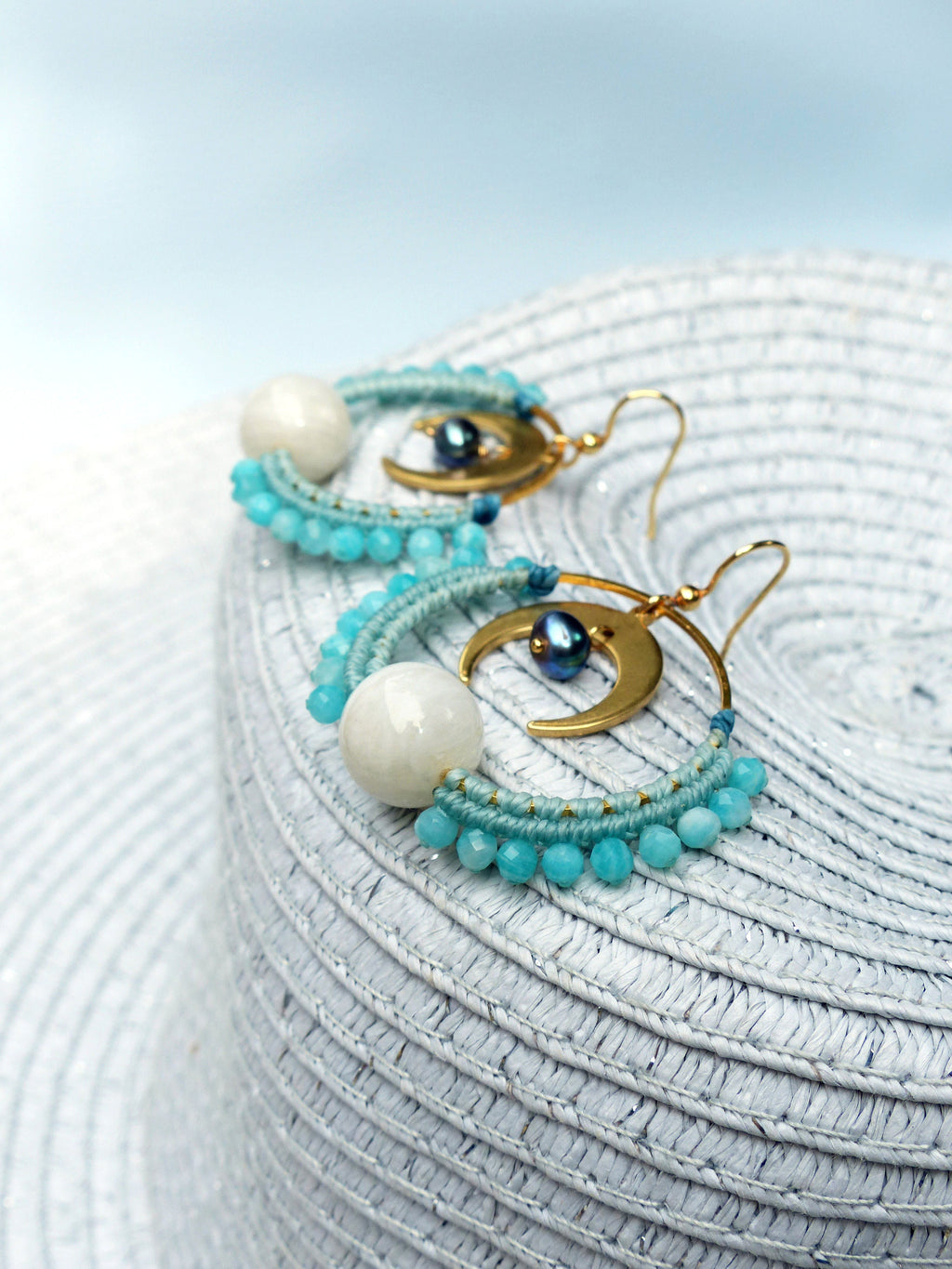 Peut inclure: Une paire de boucles d'oreilles créoles dorées avec un charme de croissant de lune et des perles bleues et blanches. Les boucles d'oreilles sont sur un chapeau de paille blanc.