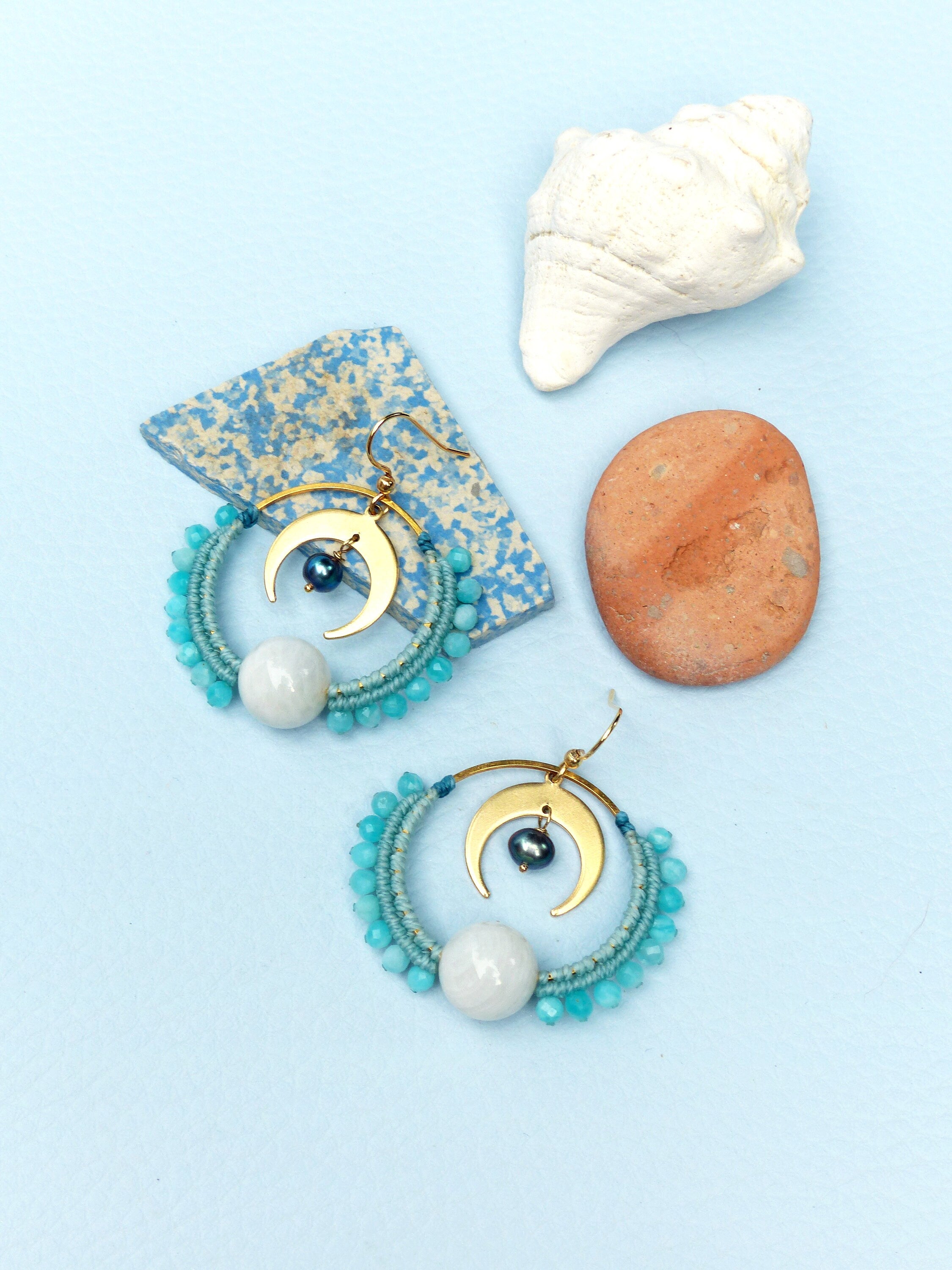 Peut inclure: Une paire de boucles d'oreilles créoles dorées avec un charme de croissant de lune et une perle de pierre blanche. Les boucles d'oreilles sont faites de perles bleues et turquoise.