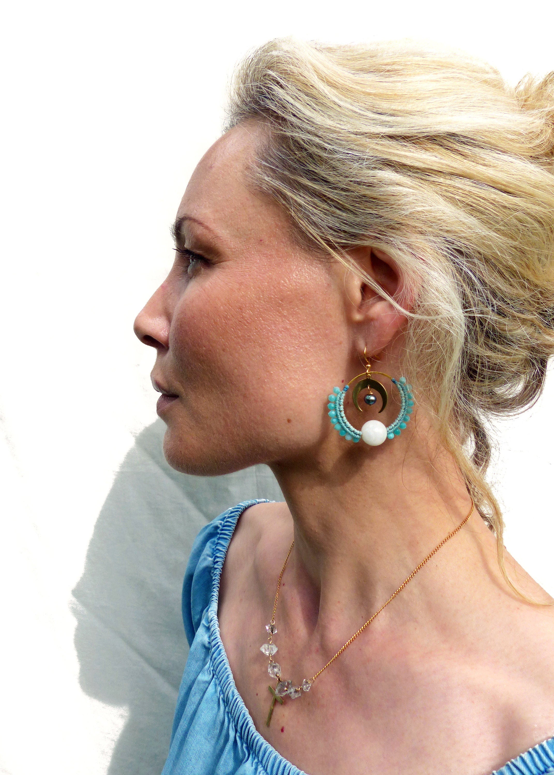 Peut inclure: Une femme portant un haut bleu clair à épaules dénudées avec un collier en or orné d'une croix en cristal et de pierres claires. Elle porte de grandes boucles d'oreilles créoles dorées avec des perles turquoise et blanches.