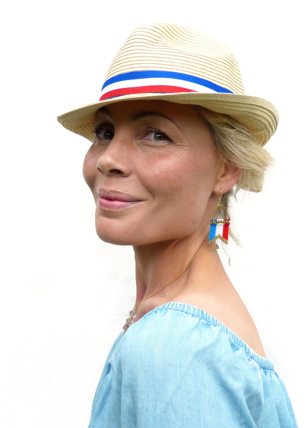 Peut inclure: Un chapeau fedora en paille avec une bande rouge, blanche et bleue. Le chapeau est porté avec un haut bleu clair à épaules dénudées et des boucles d'oreilles perlées avec des accents rouges, blancs et bleus. Un collier en or est également visible.