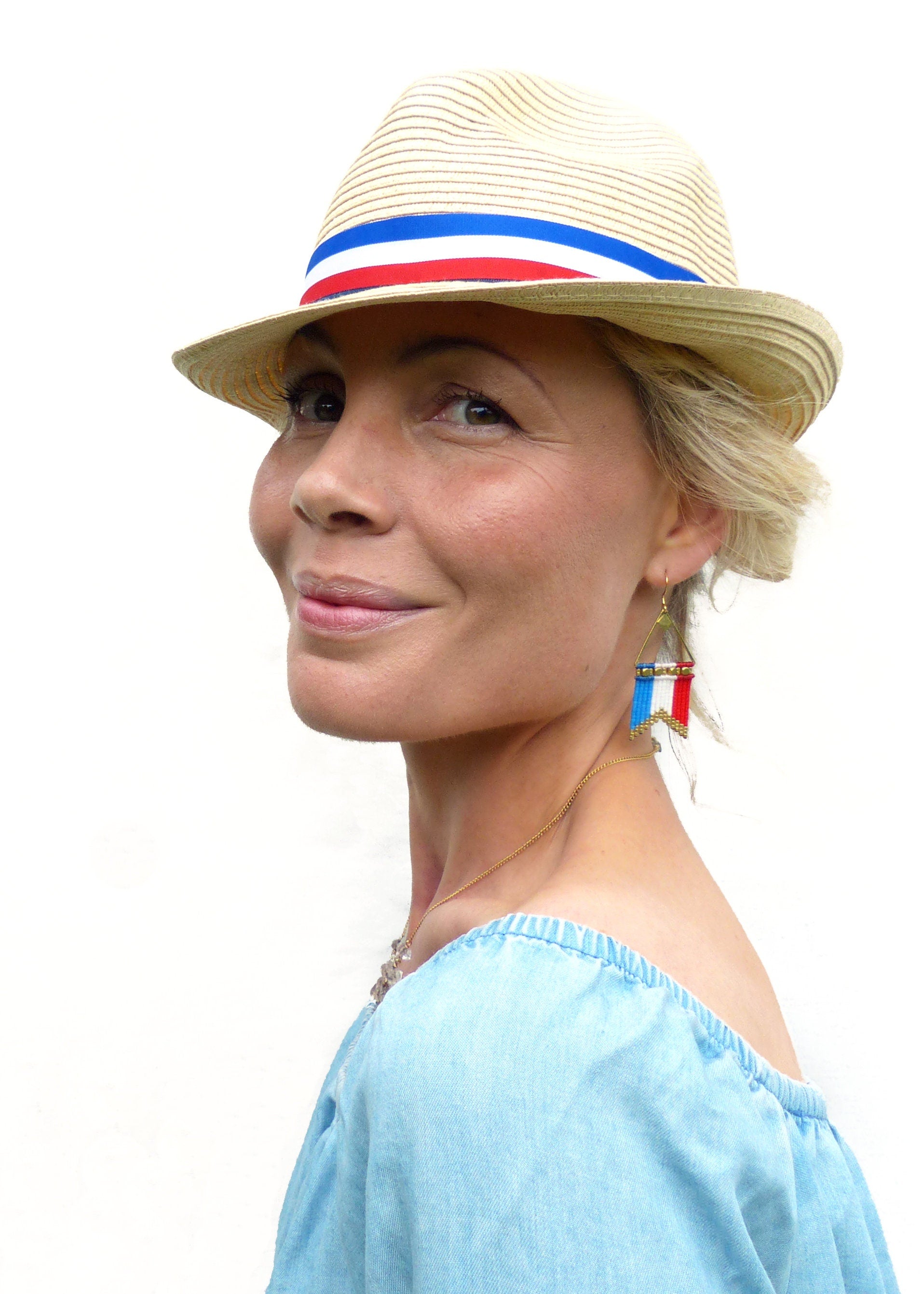 Peut inclure: Un chapeau fedora en paille avec une bande rouge, blanche et bleue. Le chapeau est porté avec un haut bleu clair à épaules dénudées et des boucles d'oreilles perlées avec des accents rouges, blancs et bleus. Un collier en or est également visible.