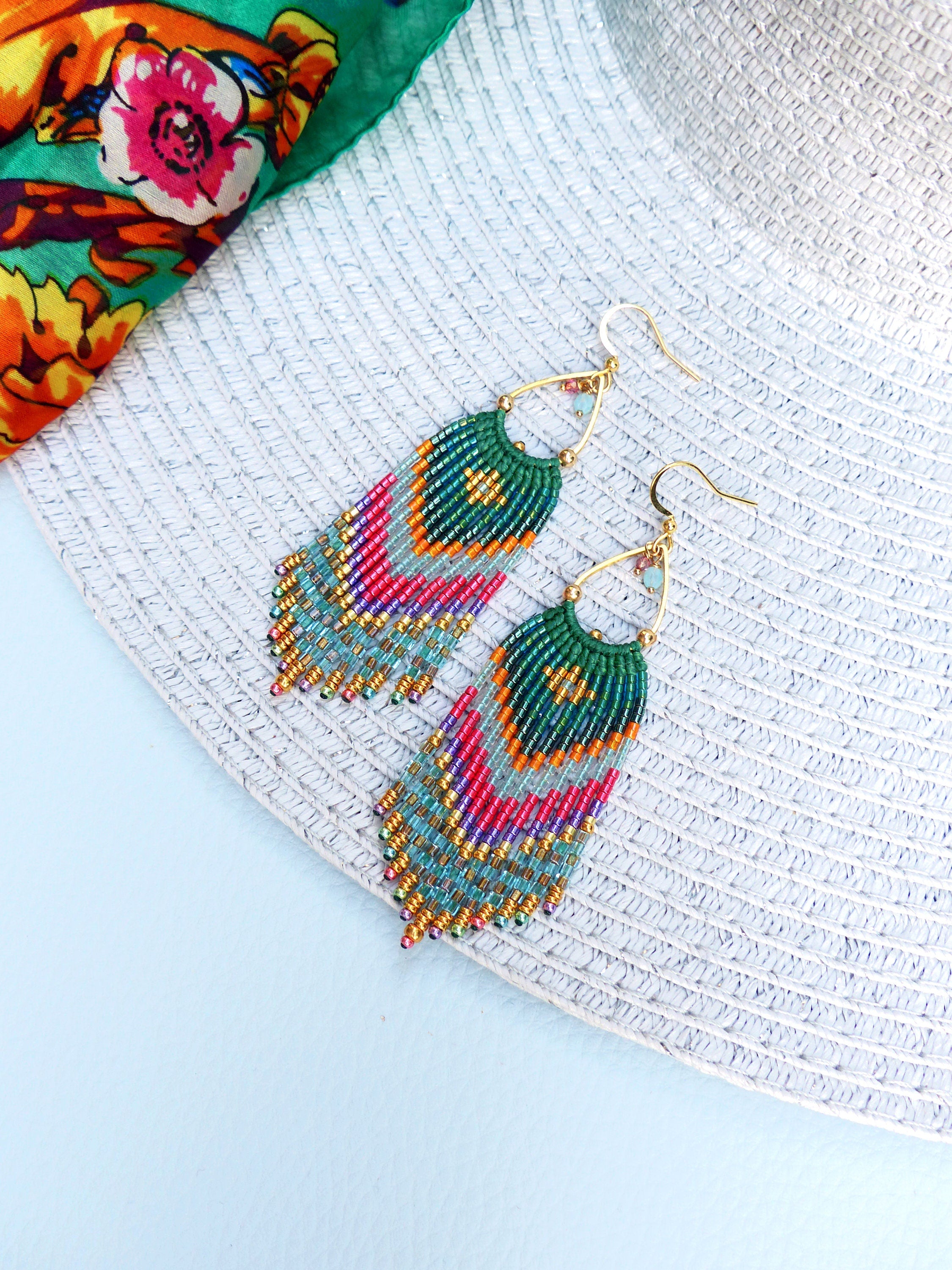 Peut inclure: Une paire de longues boucles d'oreilles en perles colorées avec un crochet doré. Les boucles d'oreilles sont faites avec une variété de perles, notamment turquoise, vert, rose et or. Les boucles d'oreilles sont suspendues à une surface blanche.