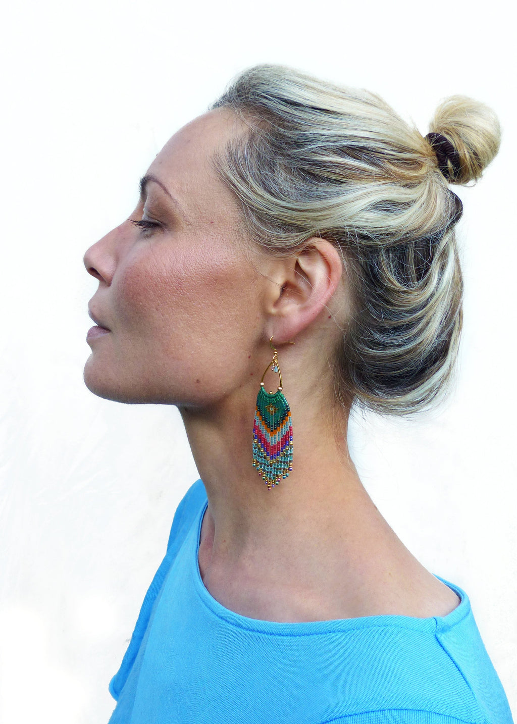 Peut inclure: Une femme aux cheveux blonds en chignon porte une paire de longues boucles d'oreilles en perles colorées. Les boucles d'oreilles sont vertes, bleues, roses et dorées. La femme porte une chemise bleu clair.
