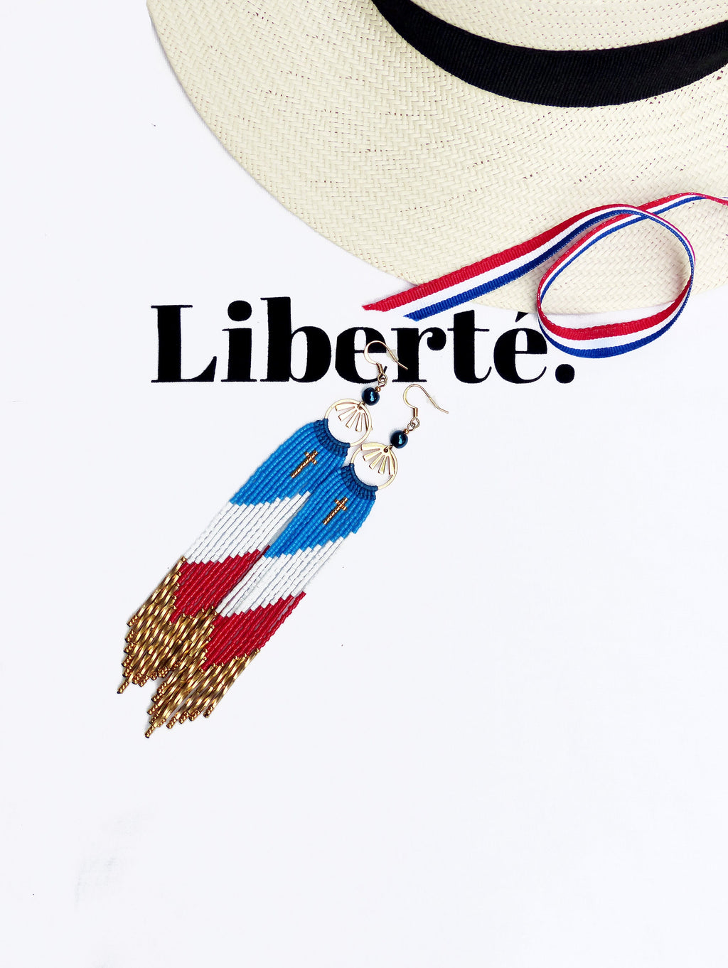 Peut inclure: Une paire de longues boucles d'oreilles en perles avec un motif rouge, blanc et bleu, rappelant le drapeau français. Les boucles d'oreilles sont suspendues à des crochets dorés et ont un petit charme en forme d'éventail doré en haut. Le mot "Liberté." est écrit en noir sur fond blanc.