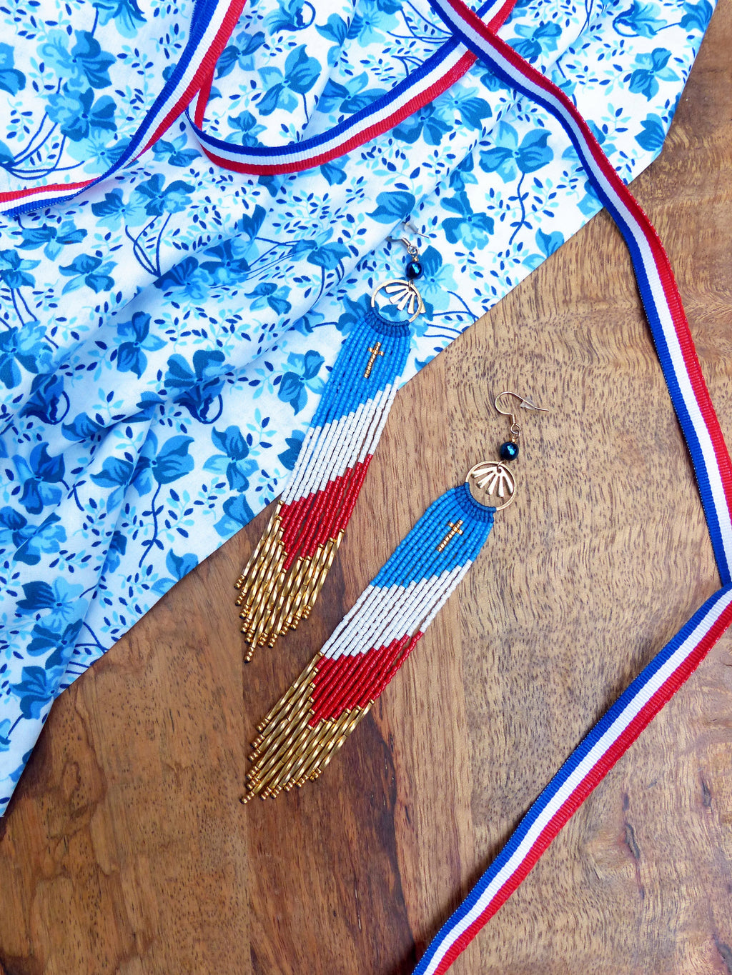 Peut inclure: Une paire de longues boucles d'oreilles en perles avec un motif rouge, blanc et bleu. Les boucles d'oreilles sont suspendues à des crochets dorés et ont un petit charme en forme de croix en haut. Les boucles d'oreilles sont sur une surface en bois avec un tissu floral bleu et blanc et un ruban rouge, blanc et bleu.