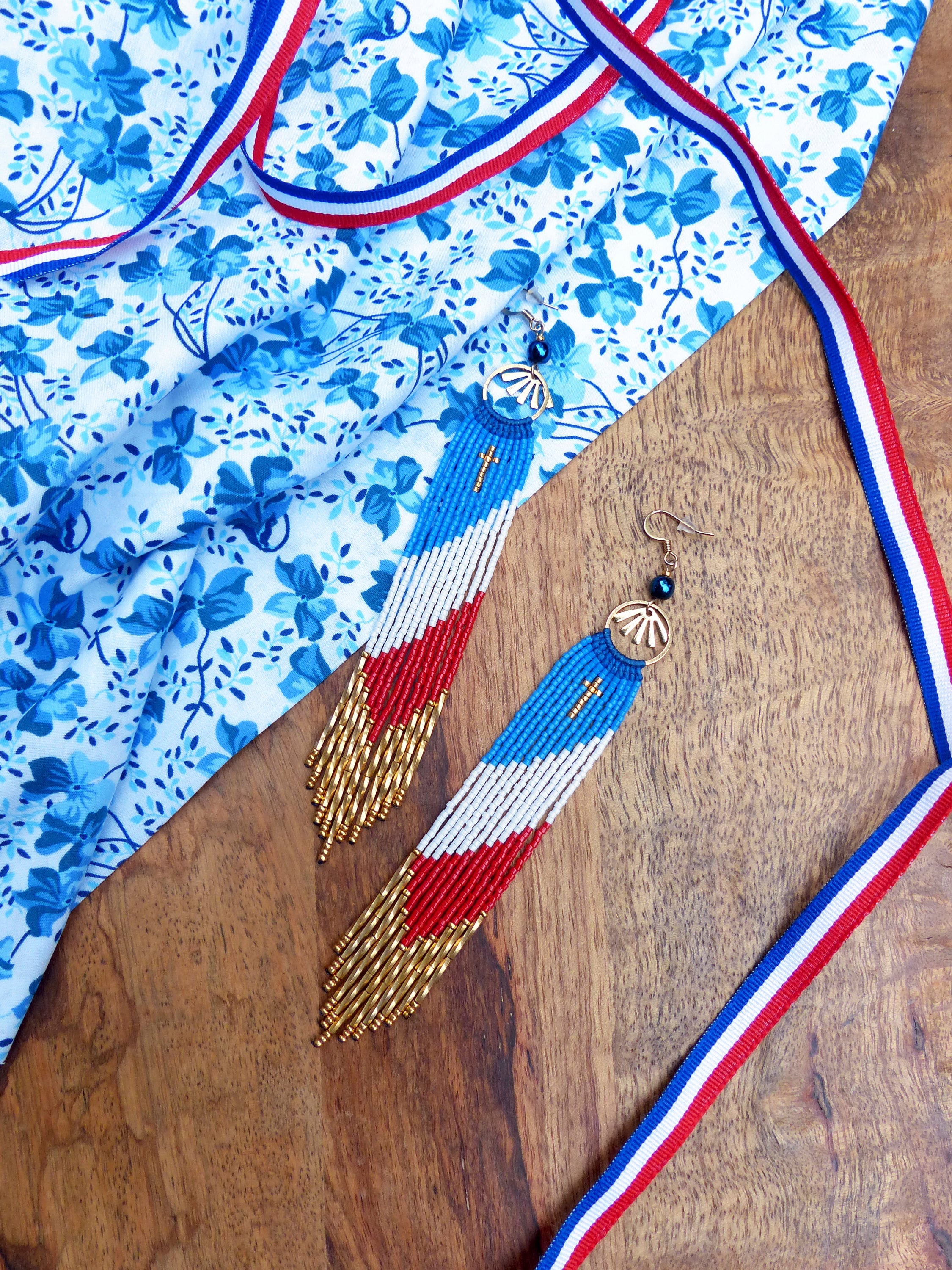 Peut inclure: Une paire de longues boucles d'oreilles en perles avec un motif rouge, blanc et bleu. Les boucles d'oreilles sont suspendues à des crochets dorés et ont un petit charme en forme de croix en haut. Les boucles d'oreilles sont sur une surface en bois avec un tissu floral bleu et blanc et un ruban rouge, blanc et bleu.