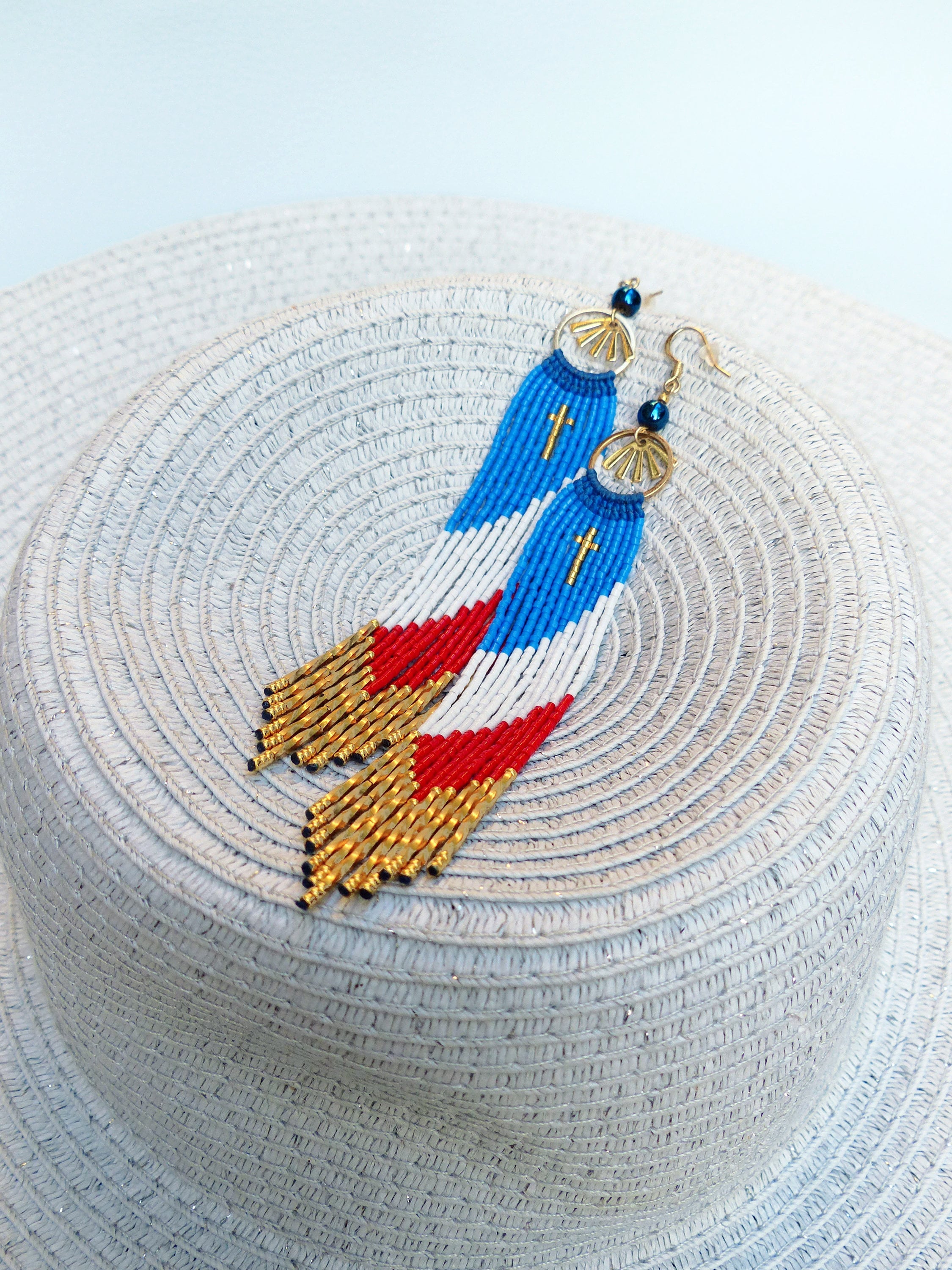Peut inclure: Une paire de longues boucles d'oreilles en perles avec un motif rouge, blanc et bleu. Les boucles d'oreilles sont ornées de perles dorées et de petites croix.