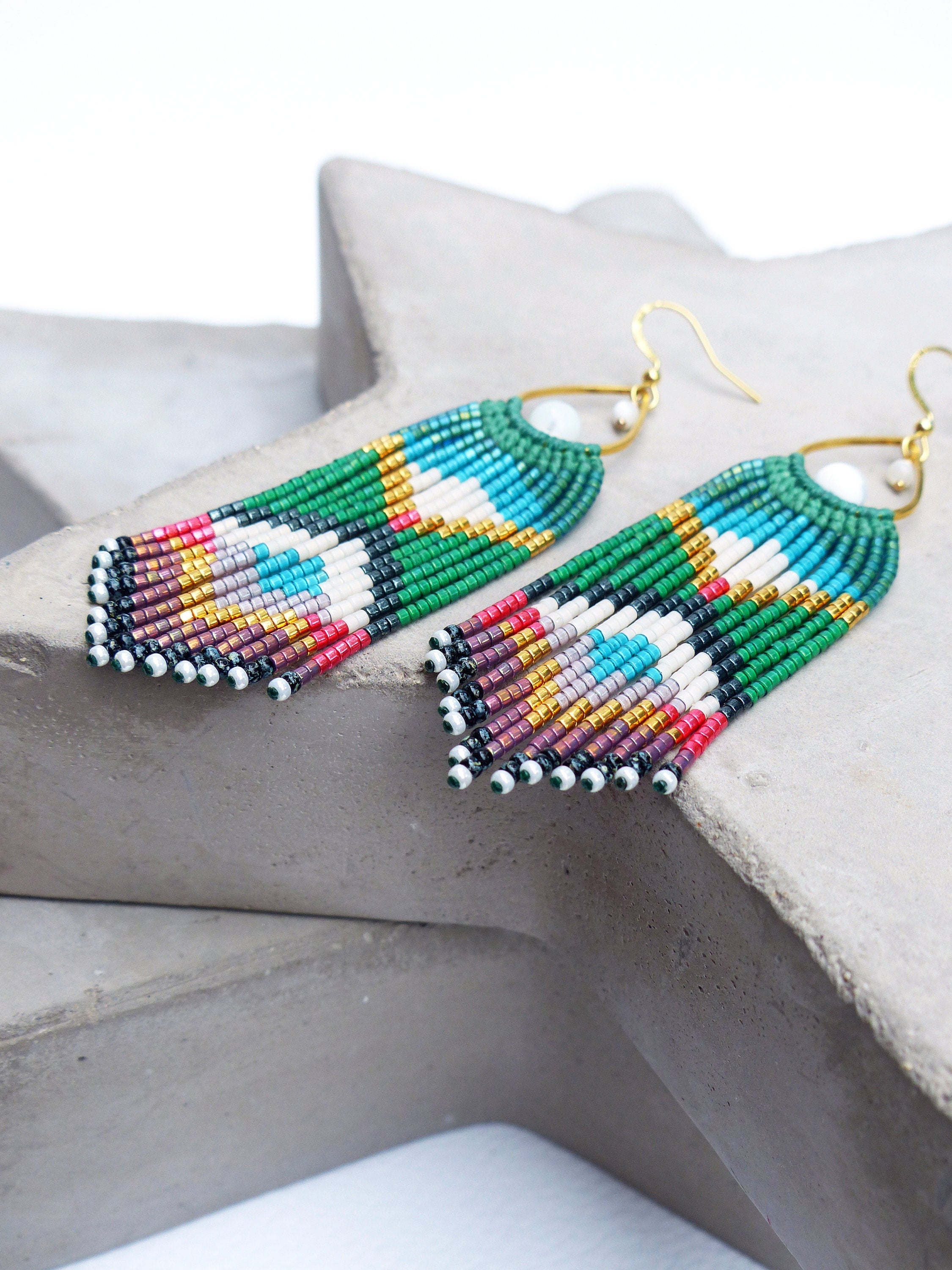 Peut inclure: Une paire de longues boucles d'oreilles en perles colorées avec une perle blanche au sommet. Les boucles d'oreilles sont faites de perles vertes, bleues, rouges, dorées et noires.