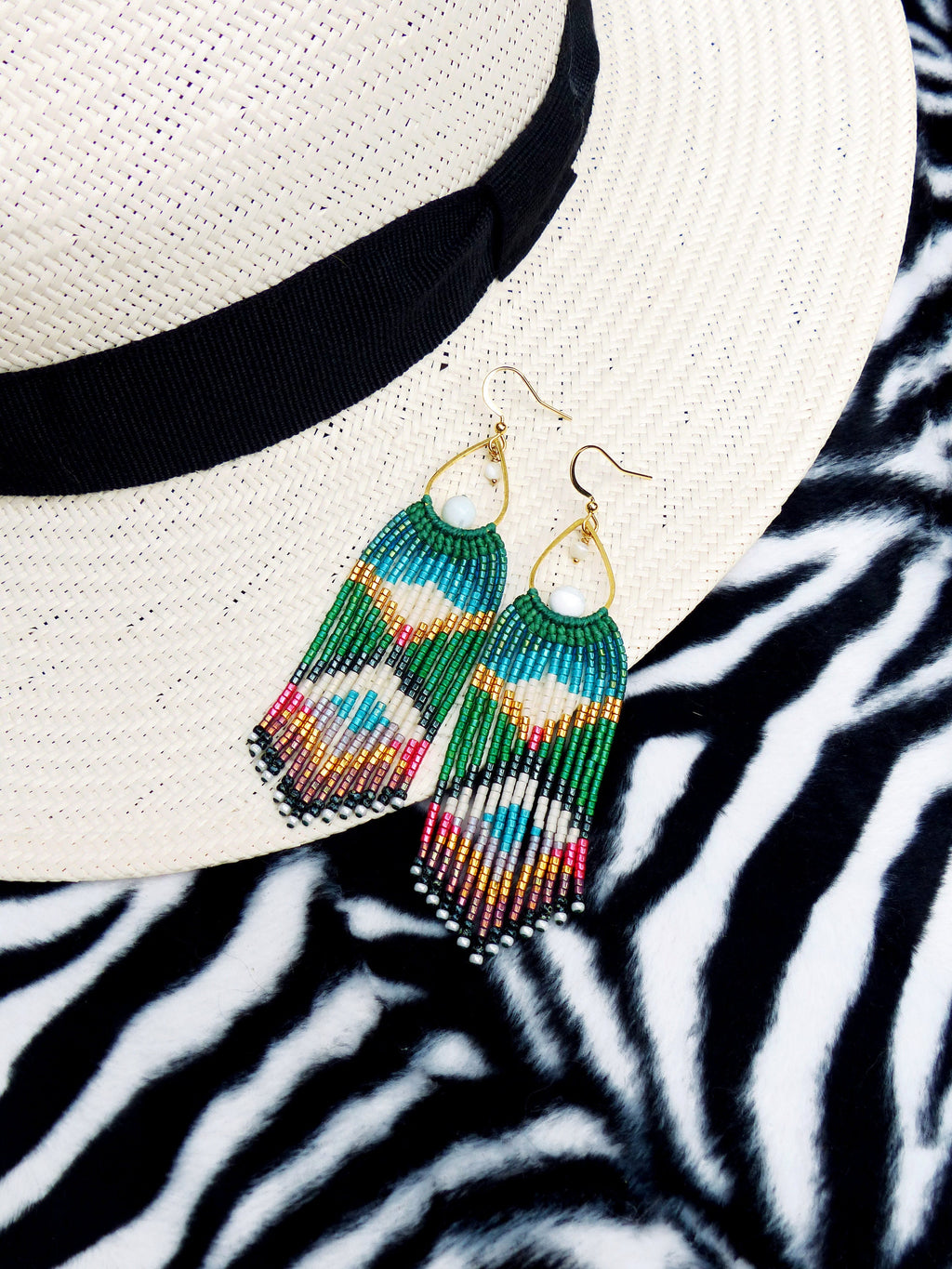 Peut inclure: Une paire de longues boucles d'oreilles en perles avec un motif vert, bleu et blanc. Les boucles d'oreilles pendent d'un chapeau de paille sur un fond à imprimé zèbre.
