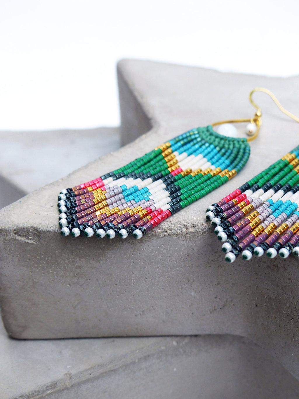 Peut inclure: Une paire de longues boucles d'oreilles en perles avec un motif géométrique dans des tons de vert, blanc, rouge, bleu, or et violet. Les boucles d'oreilles sont faites de petites perles rondes et ont une fermeture à crochet dorée.
