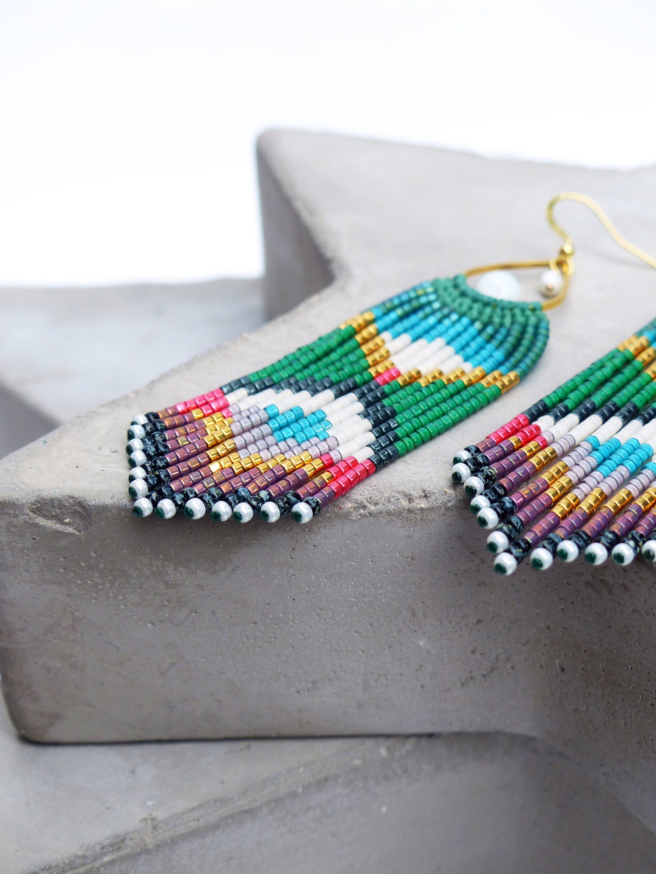 Peut inclure: Une paire de longues boucles d'oreilles en perles avec un motif géométrique dans des tons de vert, blanc, rouge, bleu, or et violet. Les boucles d'oreilles sont faites de petites perles rondes et ont une fermeture à crochet dorée.
