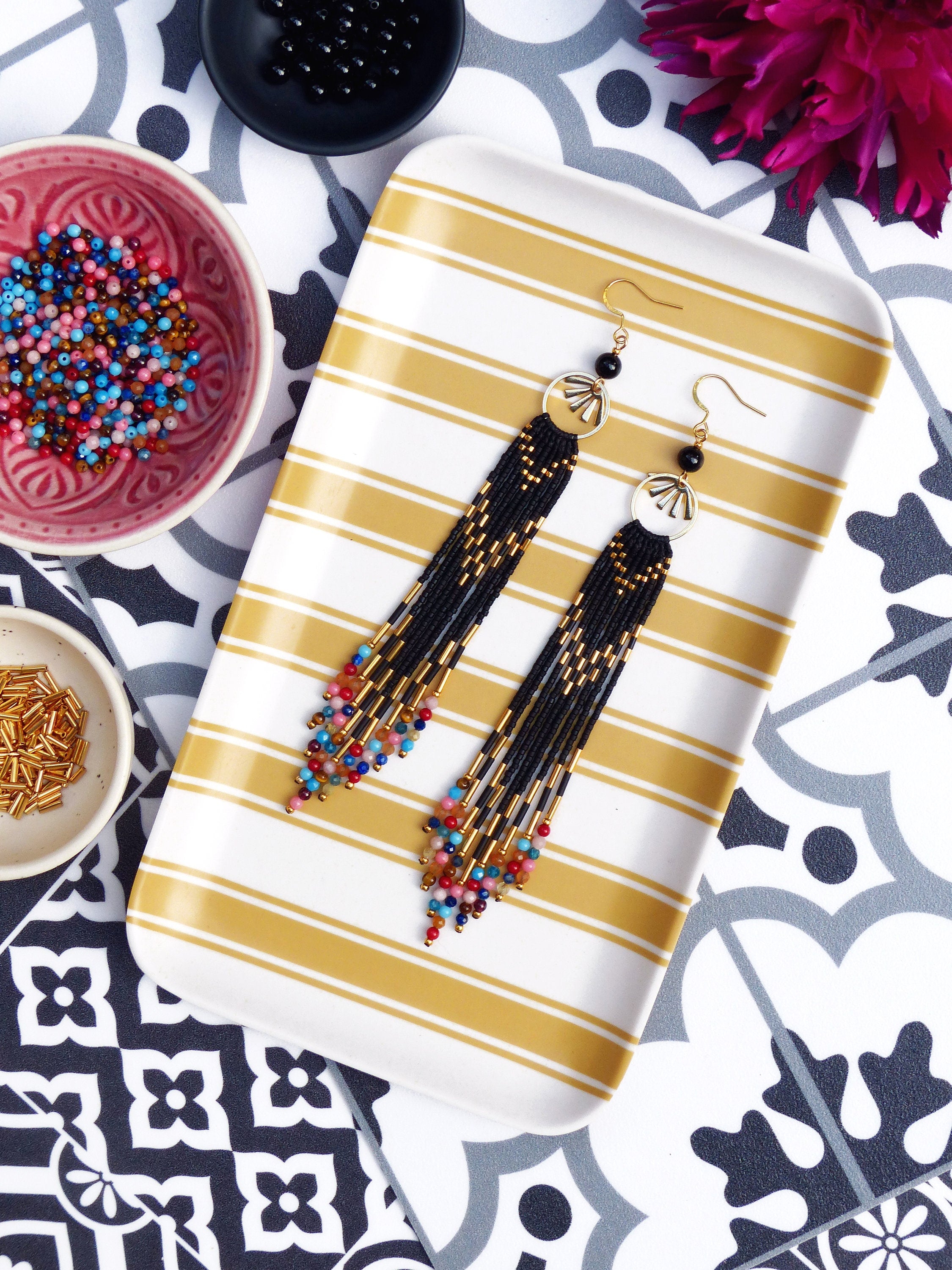 Peut inclure: Une paire de longues boucles d'oreilles noires en perles avec des accents dorés et des perles colorées. Les boucles d'oreilles sont suspendues à un plateau rayé.