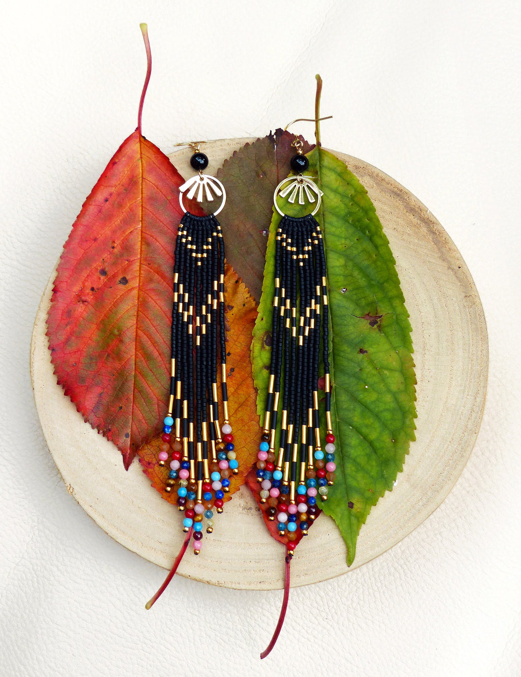 Peut inclure: Une paire de longues boucles d'oreilles en perles noires avec des accents dorés et des perles colorées qui pendent du bas. Les boucles d'oreilles sont présentées sur une assiette en bois avec des feuilles d'automne.