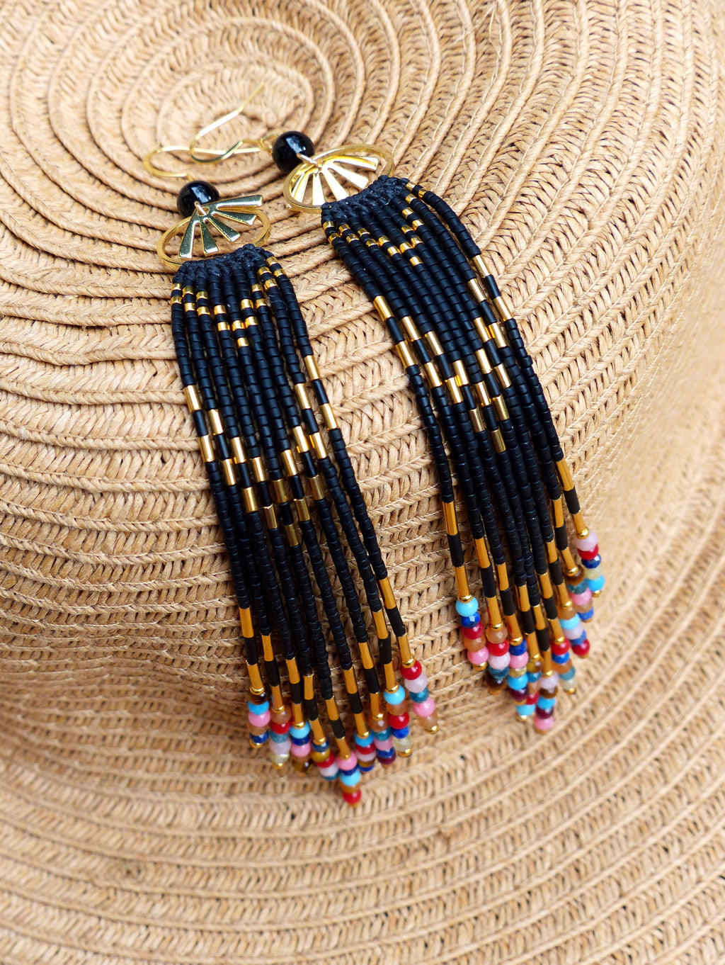 Peut inclure: Une paire de longues boucles d'oreilles en perles noires et dorées avec des accents colorés. Les boucles d'oreilles présentent un motif en forme de soleil en haut et pendent jusqu'en bas.