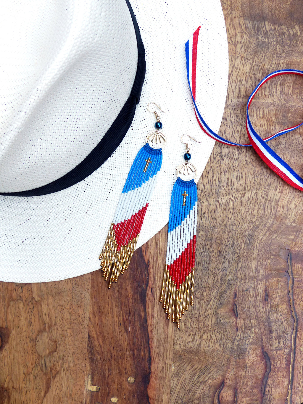 Peut inclure: Une paire de longues boucles d'oreilles en perles avec un motif bleu, blanc et rouge. Les boucles d'oreilles ont un pompon doré et un petit charme en forme de croix. Les boucles d'oreilles sont présentées sur une surface en bois avec un chapeau blanc et un ruban rouge, blanc et bleu.