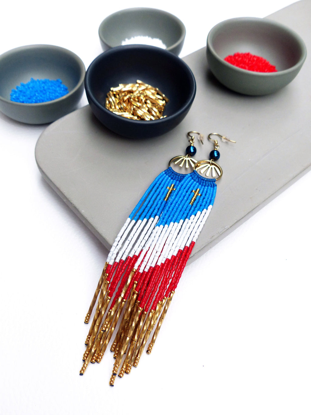 Peut inclure: Une paire de longues boucles d'oreilles en perles avec un motif rouge, blanc et bleu. Les boucles d'oreilles présentent un motif en éventail doré en haut et pendent avec une frange de perles dorées.