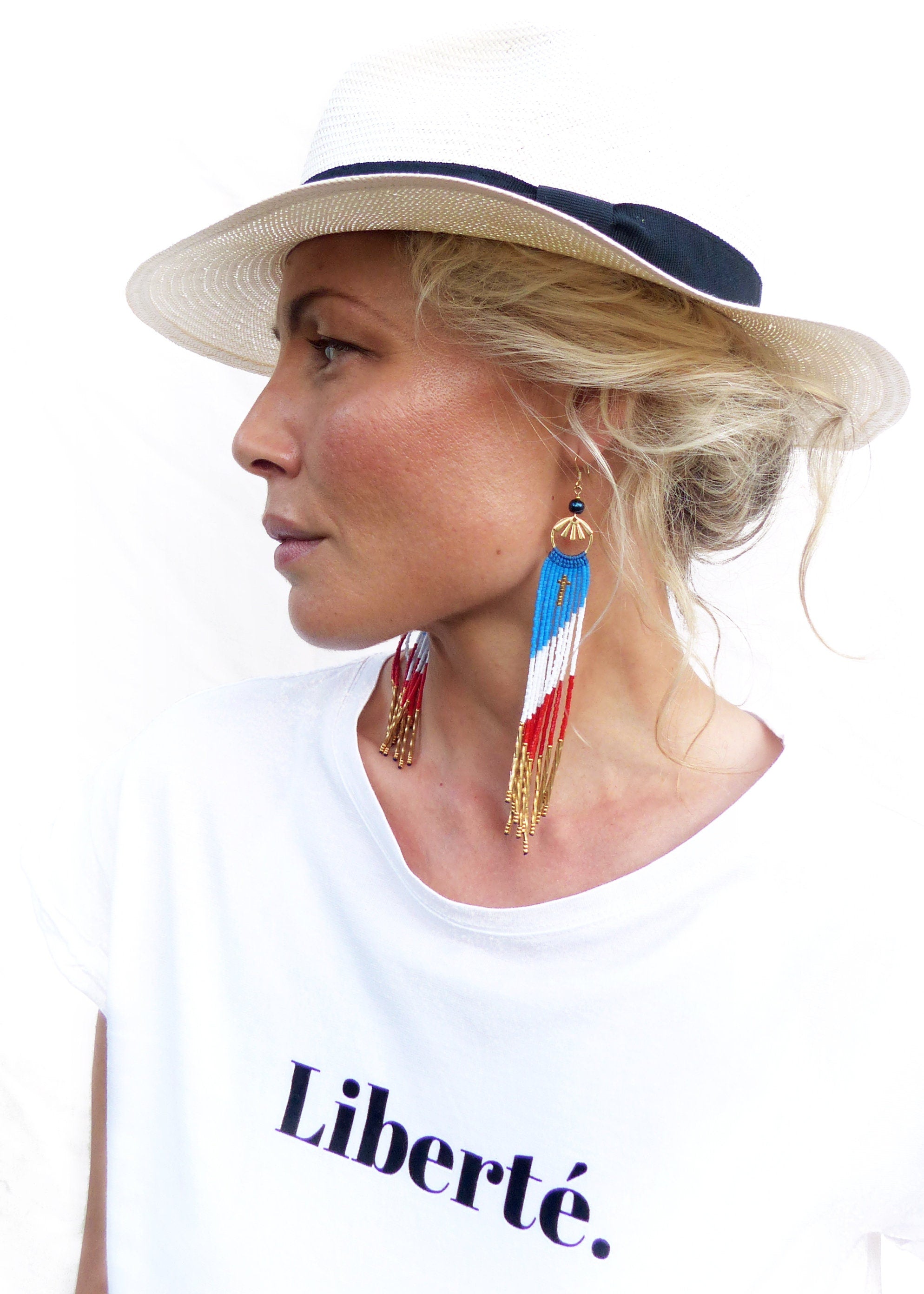 Peut inclure: Une femme portant un chapeau de paille blanc avec un bandeau noir et un t-shirt blanc sur lequel est imprimé le mot "Liberté". Elle porte de longues boucles d'oreilles en perles de couleurs rouge, blanche et bleue.
