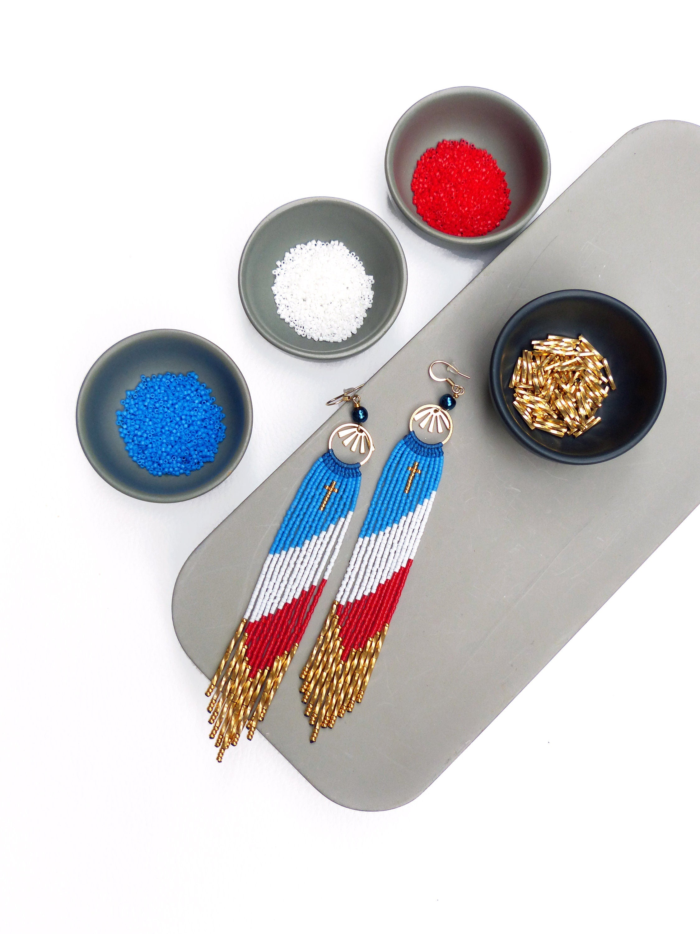 Peut inclure: Une paire de longues boucles d'oreilles en perles avec un motif rouge, blanc et bleu. Les boucles d'oreilles sont suspendues sur une surface grise avec de petits bols de perles rouges, blanches et bleues.