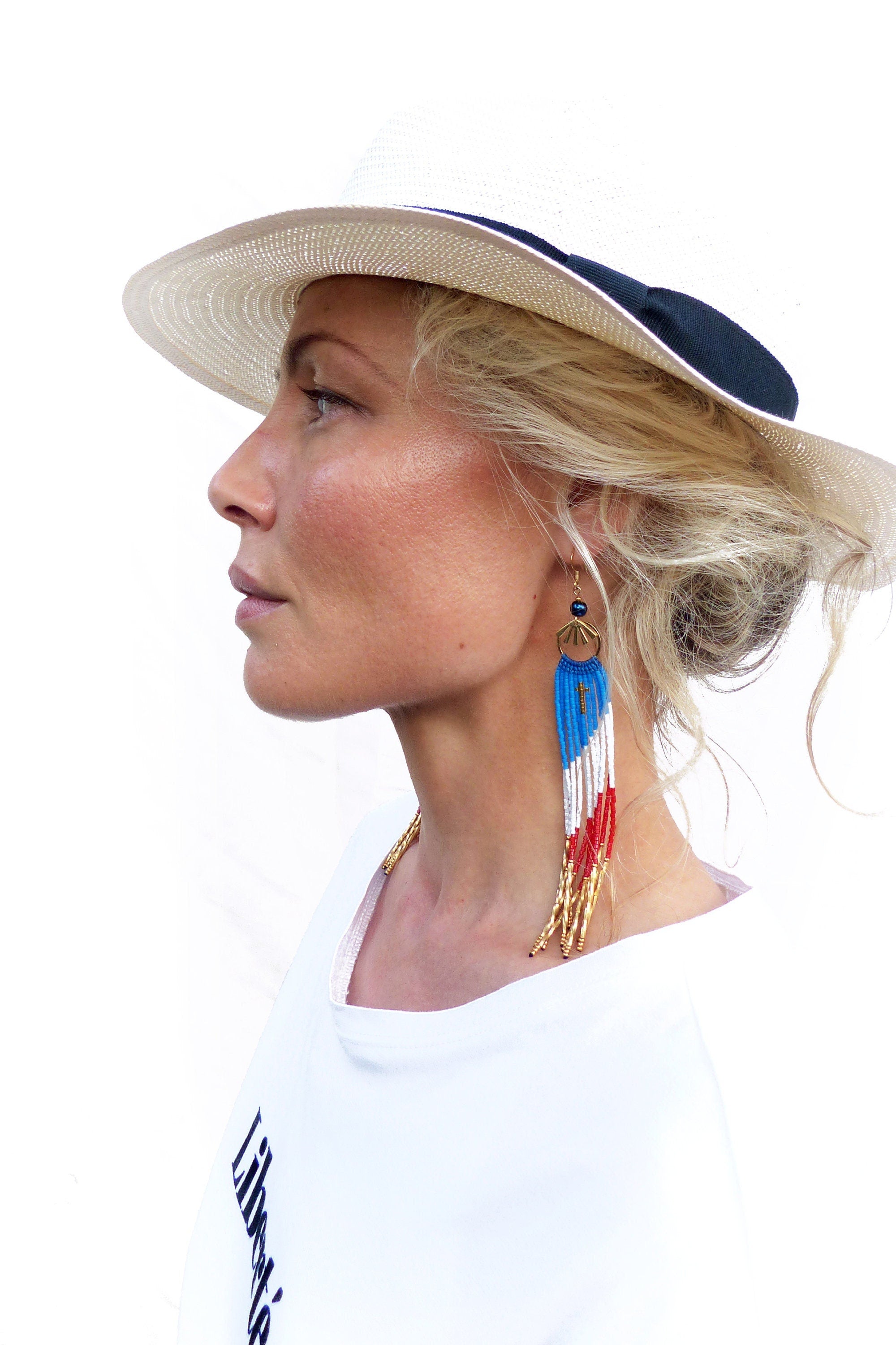 Peut inclure: Une femme portant un chapeau blanc avec un bandeau noir et un t-shirt blanc avec le mot "Liberty" imprimé dessus. Elle porte de longues boucles d'oreilles en perles avec des perles rouges, blanches et bleues.