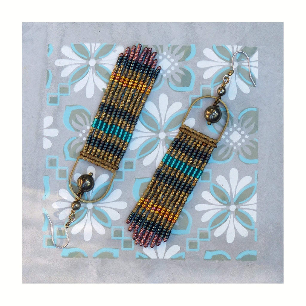 Peut inclure: Une paire de longues boucles d'oreilles en perles avec un motif géométrique dans des tons d'or, de brun, de turquoise et de rose. Les boucles d'oreilles sont suspendues à un anneau en laiton avec une perle gris foncé.