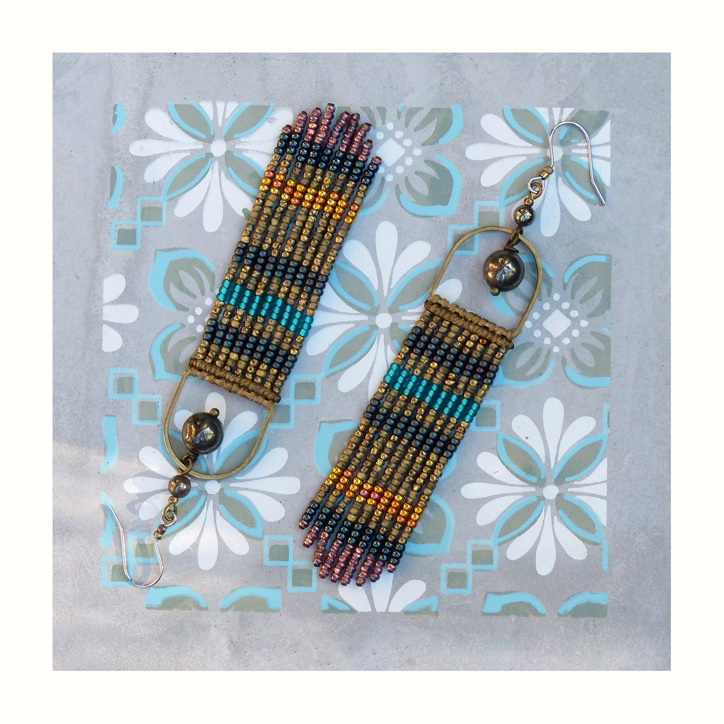 Peut inclure: Une paire de longues boucles d'oreilles en perles avec un motif géométrique dans des tons d'or, de brun, de turquoise et de rose. Les boucles d'oreilles sont suspendues à un anneau en laiton avec une perle gris foncé.