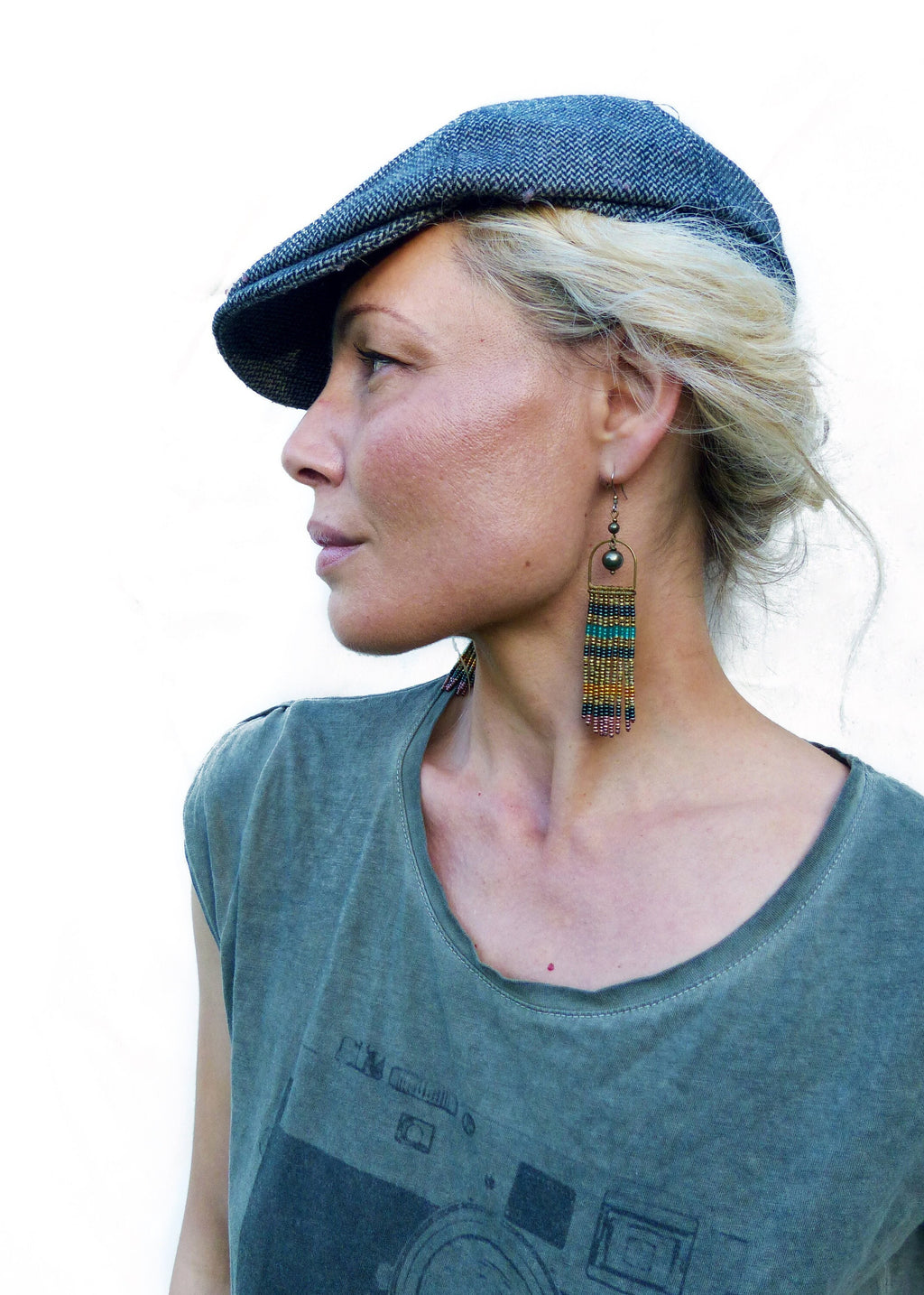 Peut inclure: Une femme portant une casquette de newsboy bleue et un t-shirt vert avec un motif de caméra. Elle porte de longues boucles d'oreilles en perles colorées.
