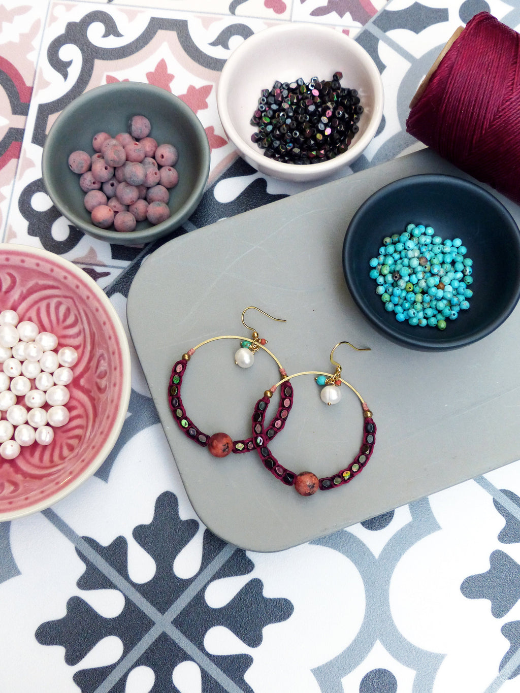 Peut inclure: Une paire de boucles d'oreilles créoles avec un cordon de velours bordeaux et des perles dorées. Les boucles d'oreilles sont sur une surface grise avec de petits bols de perles en arrière-plan.