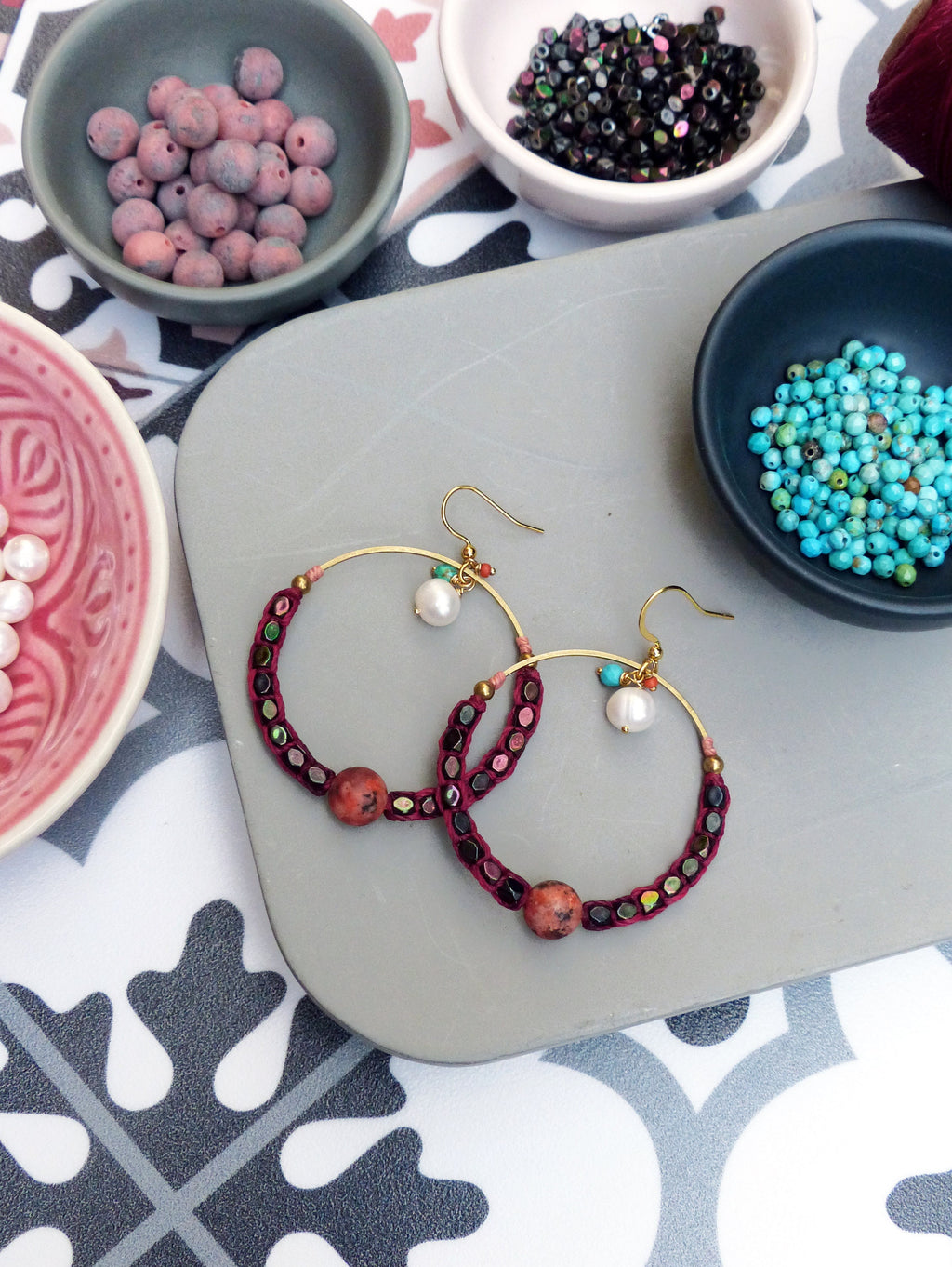 Peut inclure: Une paire de boucles d'oreilles créoles dorées avec un tissu bordeaux enroulé autour du cercle. Les boucles d'oreilles sont décorées de petites perles colorées et d'une perle ronde plus grande au bas de chaque cercle.