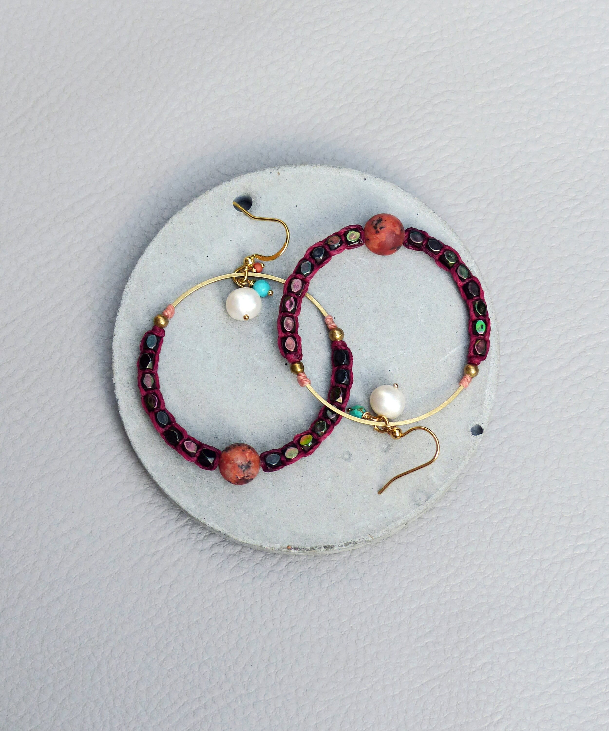 Peut inclure: Une paire de boucles d'oreilles créoles avec un design de perles bordeaux et noires. Les boucles d'oreilles ont un anneau en fil d'or et un petit charme en perle.