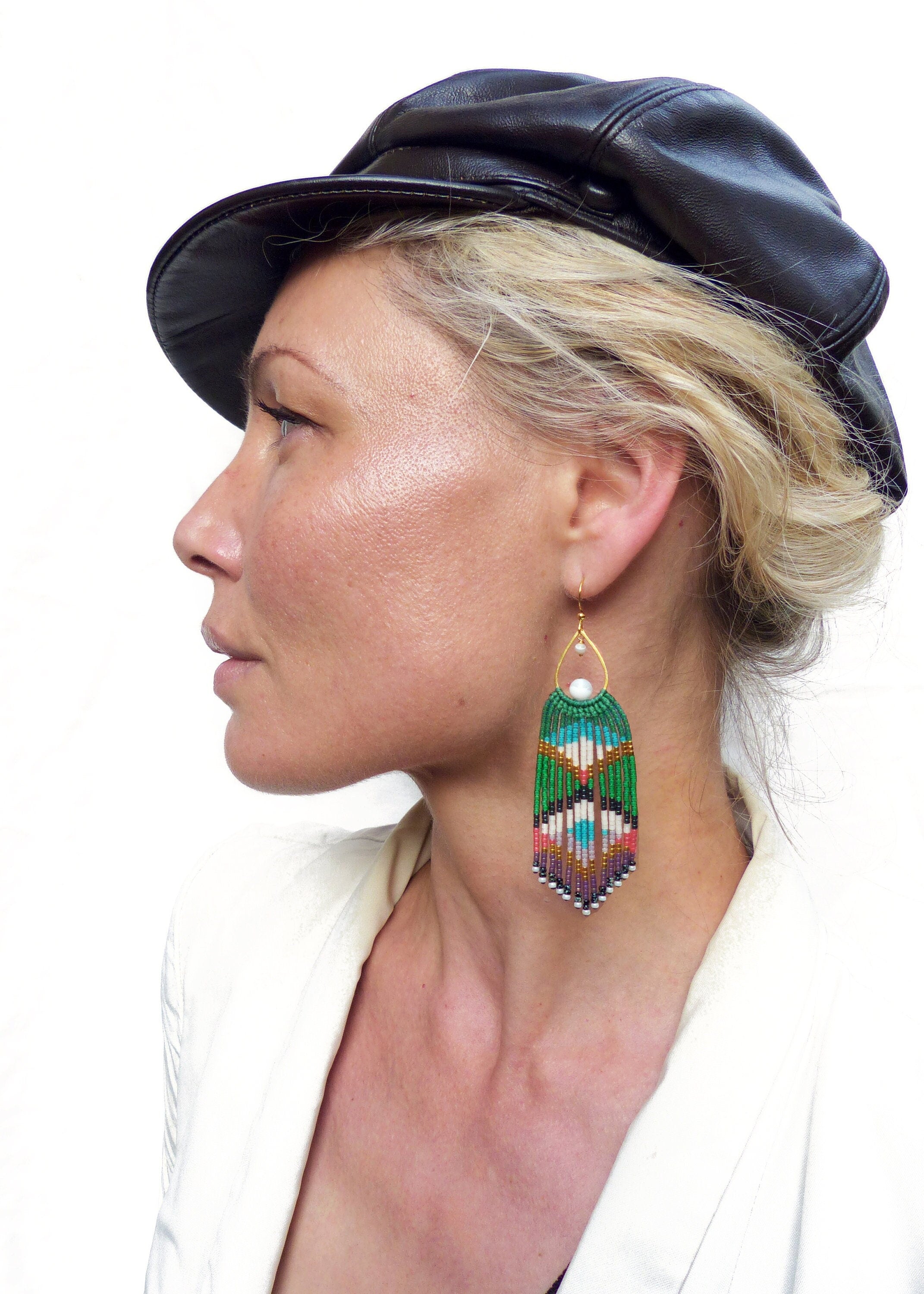 Peut inclure: Une femme portant une casquette en cuir noir et un blazer blanc avec de longues boucles d'oreilles en perles vertes et multicolores.