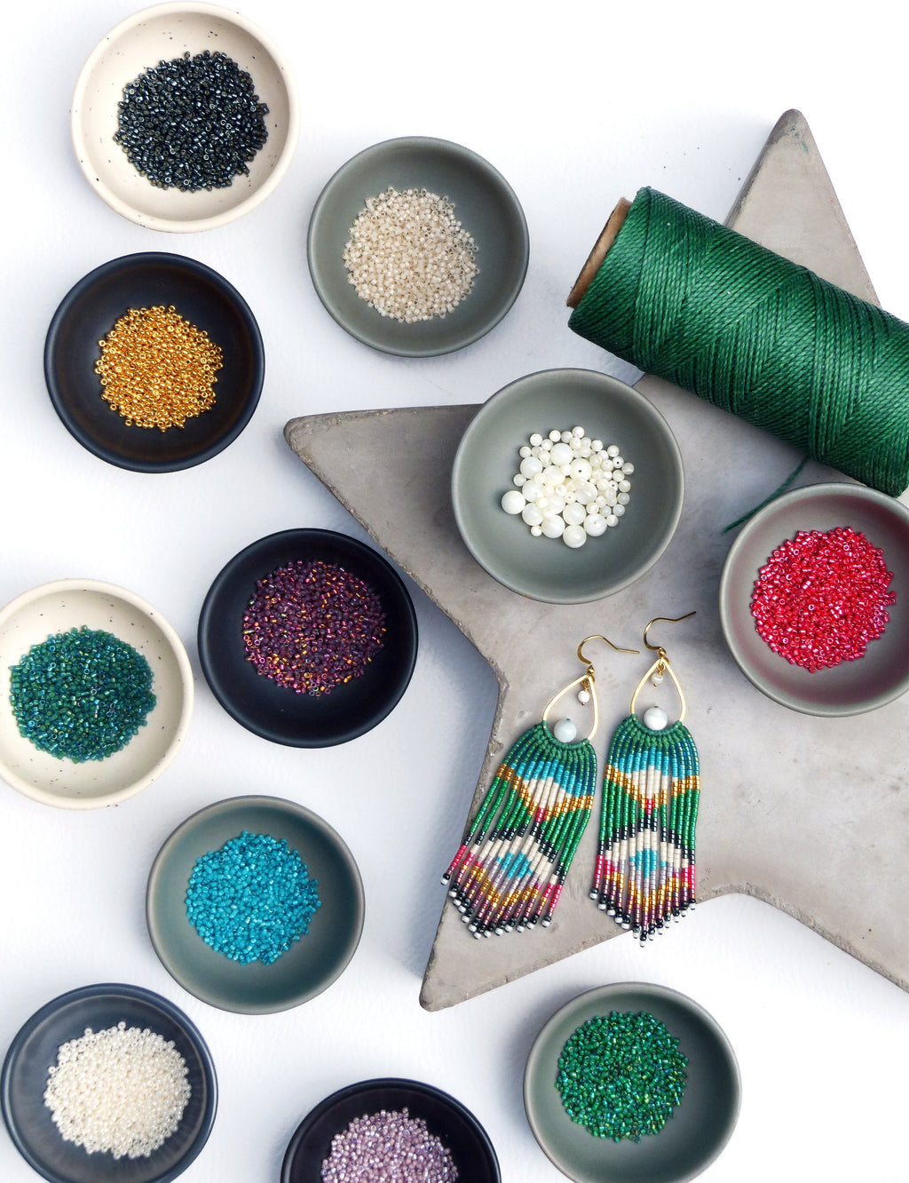Peut inclure: Une paire de longues boucles d'oreilles en perles colorées avec un motif géométrique vert, blanc et bleu. Les boucles d'oreilles sont présentées sur une surface en forme d'étoile grise avec divers petits bols de perles de différentes couleurs, notamment noir, or, blanc, rouge, violet, bleu sarcelle et vert.
