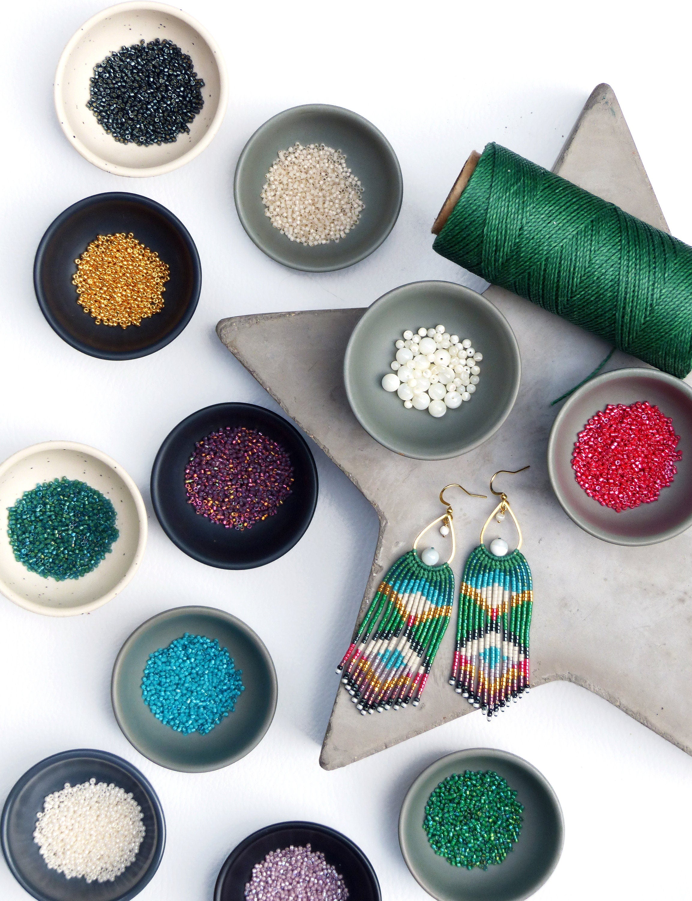 Peut inclure: Une paire de longues boucles d'oreilles en perles colorées avec un motif géométrique vert, blanc et bleu. Les boucles d'oreilles sont présentées sur une surface en forme d'étoile grise avec divers petits bols de perles de différentes couleurs, notamment noir, or, blanc, rouge, violet, bleu sarcelle et vert.